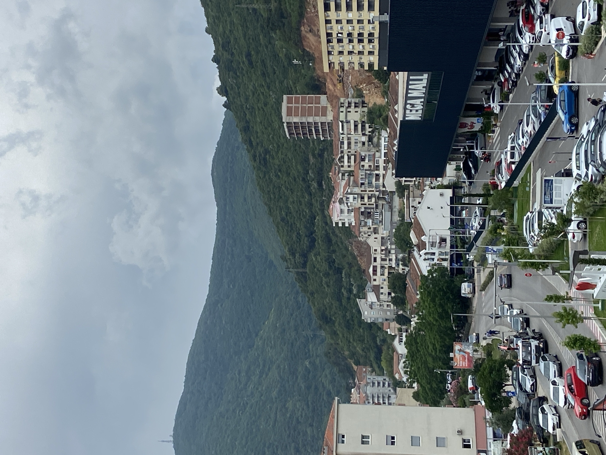 This image captures an elevated view of Budva, Montenegro, showcasing an urban landscape at the base of a large, forested mountain range under an overcast sky.
On the left side of the frame, a sprawling, dark green mountain dominates, its slopes densely covered in trees, receding into the distance. Power lines are faintly visible against the mountain's greenery in some sections. The sky above is largely cloudy, featuring a soft, diffused light with no direct sunlight.
The right side of the image depicts a developed town area. Numerous buildings of varying sizes and architectural styles are present, ranging from multi-story apartment complexes to commercial structures. A large, dark-colored building prominently displays "MEGA MALL" in white capital letters on its facade, indicating a retail center. Around the mall and other buildings, extensive parking lots and roads are filled with a multitude of vehicles, including white, black, blue, and red cars, many of which appear to be new. Some structures are visible in various stages of construction or renovation, with exposed concrete frames.
Small, indistinct figures of people can be seen on sidewalks and near the parked cars, suggesting routine daily activities. The scene portrays a bustling town that thrives alongside a dramatic natural backdrop. The time of day appears to be daytime, given the ambient light, but the overcast conditions lend a subdued atmosphere to the entire scene.