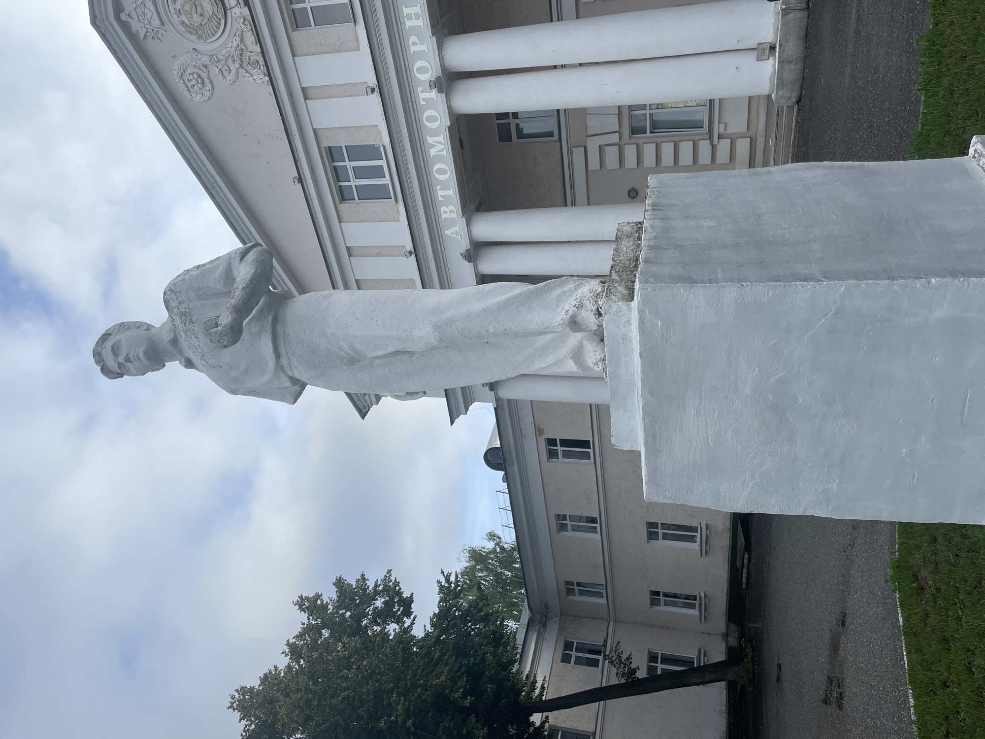 The image shows a white stone statue of a young man in work clothes standing on a white pedestal in front of a building in Zavolzhye, Russia. The statue is in the foreground, occupying the right half of the frame. The man is depicted in a dynamic pose, with his right hand raised to his chest and his left hand holding what appears to be a small object. He wears a collared shirt and loose-fitting trousers. The building behind the statue is a multi-story structure with a neoclassical facade, featuring white columns and large windows. The words "АВТОМОТОРН" are visible in large letters above the columns. To the left of the statue, a large tree with dark green foliage partially obscures the building. The ground in front of the statue is paved with asphalt and grass. The sky is overcast with clouds.