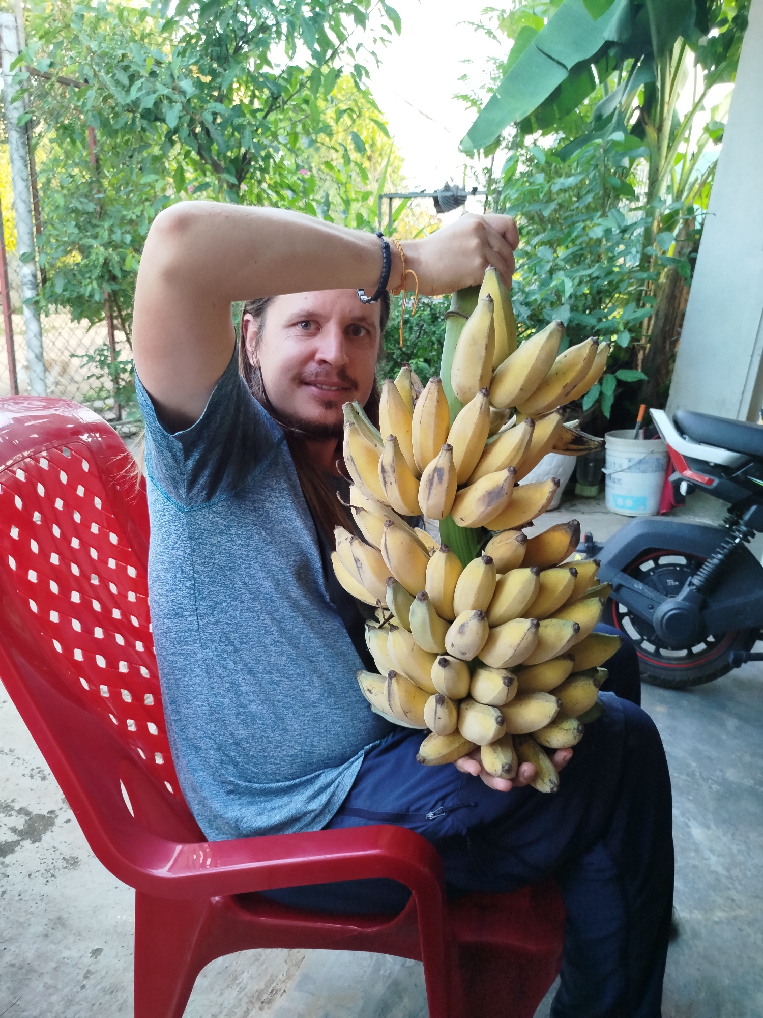 A man is sitting on a red plastic chair outdoors in Bờ Y, Vietnam. He is holding a large bunch of yellow bananas in one hand while his other arm is raised overhead. He has medium-length hair, a mustache, and a slight smile. He is wearing a gray t-shirt and dark blue pants. Behind him are trees, a fence, a building, and a parked moped. A white bucket is also visible in the background.
