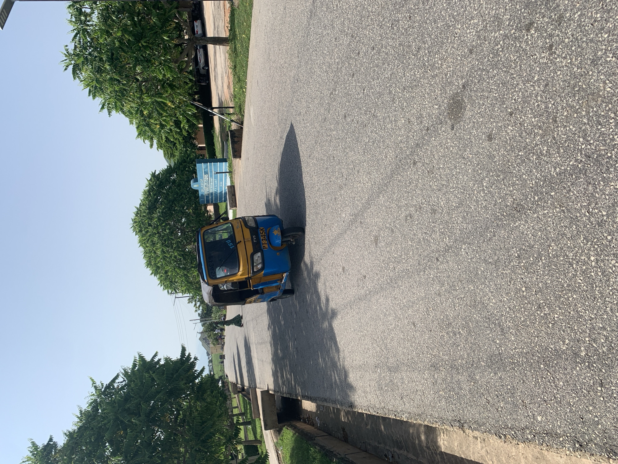 In Jalingo, Nigeria, on a paved road, a blue and yellow auto-rickshaw with the license plate PT345K is parked facing away from the viewer. The auto-rickshaw is on the right side of the road, casting a shadow on the asphalt. On the left side of the road, a concrete drainage channel runs alongside the pavement. Further down the road, several people are visible walking. To the right of the road, there is a blue sign that reads "COLLEGE OF POSTGRADUATE STUDIES, TARABA STATE UNIVERSITY". Trees line both sides of the road, and the sky is clear and blue.
