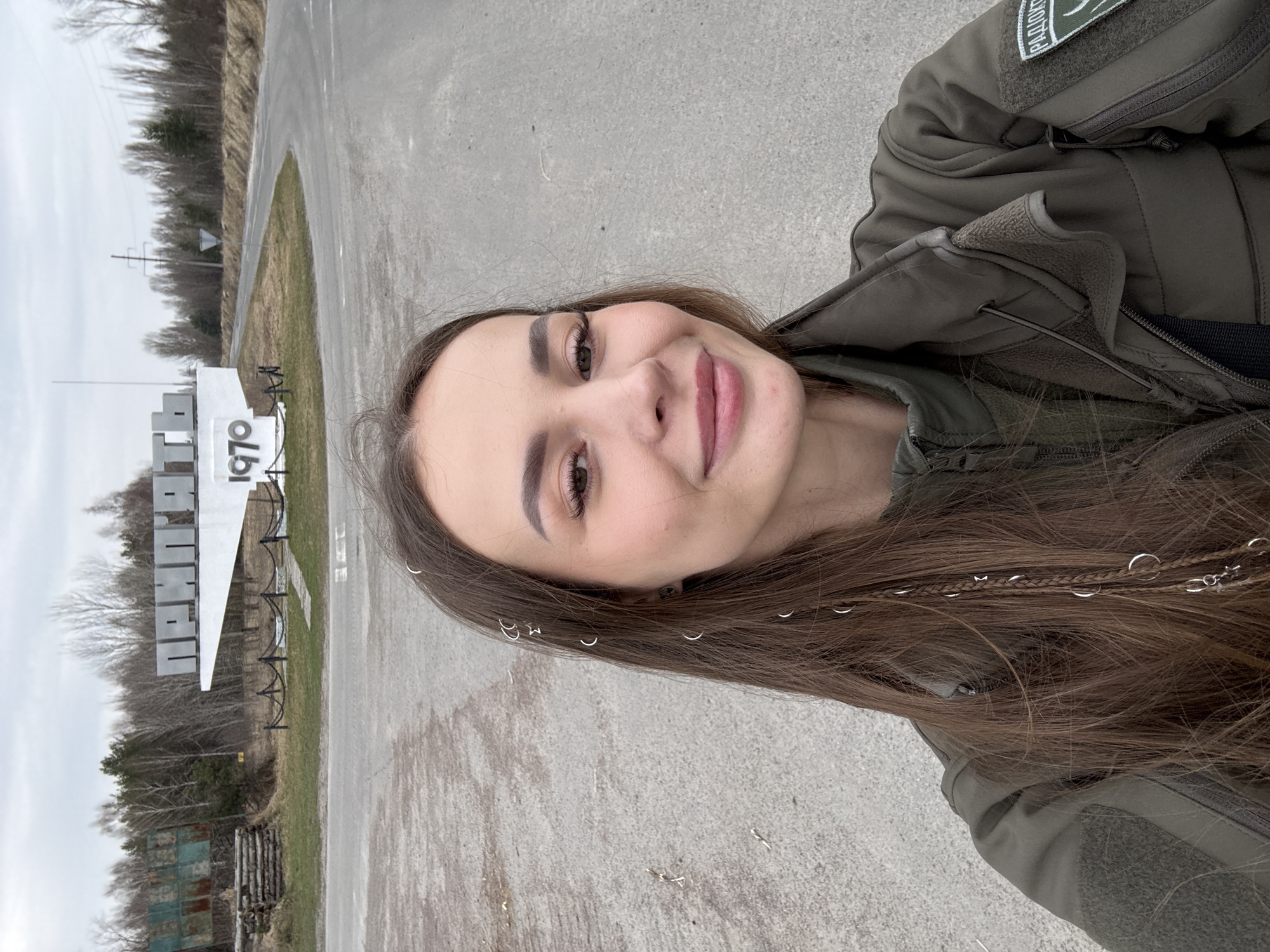 A medium shot depicts a fair-skinned, smiling woman wearing a green tactical jacket, presumably in the city of Pryp'yat', Ukraine. Her long, brown hair is adorned with silver rings. Positioned behind her and to the left is a large, white sign featuring the lettering "ПРИП'ЯТЬ 1970", indicative of the city's name and establishment year. The background includes leafless trees and an overcast sky. The ground appears to be asphalt.