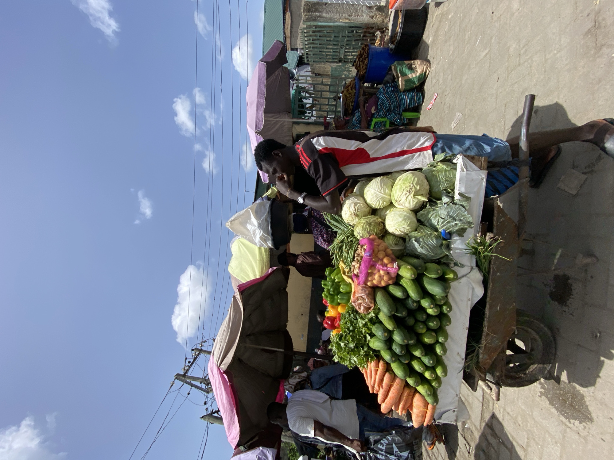 A street vendor stands behind a cart laden with fresh produce in Lekki, Nigeria. The cart displays cabbages, cucumbers, carrots, bell peppers, lettuce, and potatoes. The vendor, a dark-skinned man wearing a red and white shirt, appears to be eating. A large umbrella provides shade for the vendor and the produce. A power line is visible in the background against a blue sky with scattered clouds. Other people are seen in the area, engaged in various activities.