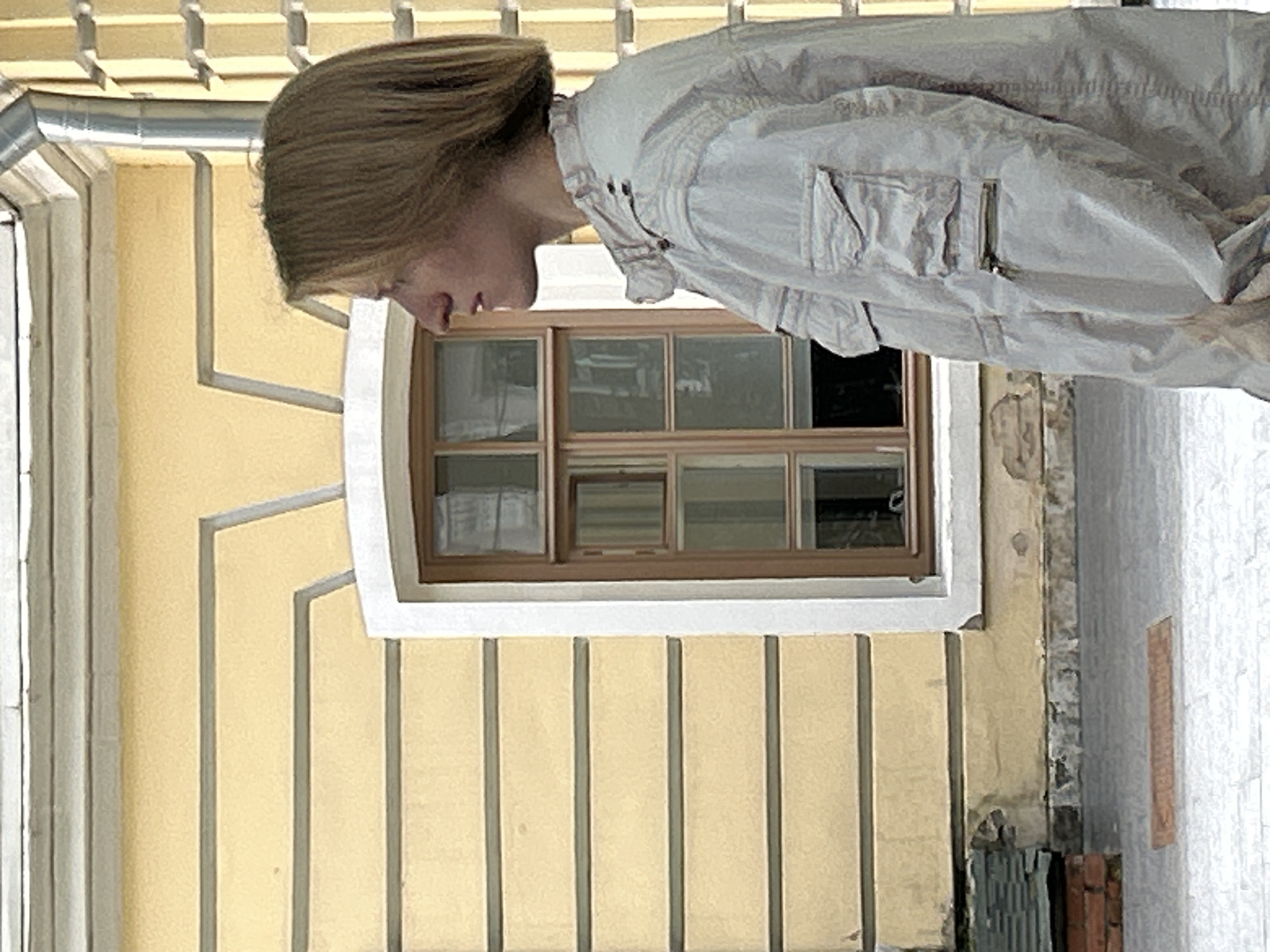 A person with light brown hair, styled with a side part and reaching to their shoulders, stands in profile facing left. They are wearing a light-colored, possibly gray or beige, jacket with multiple pockets and zippered details. The jacket has a standing collar with visible snaps. The person's nose and chin are prominent in the profile view.
They are positioned in front of a building with a light yellow facade. The facade features horizontal decorative lines in a slightly darker shade, creating a layered effect. To the left of the person is a window with a brown frame, divided into multiple panes by vertical and horizontal mullions. The window reflects some of the surrounding environment, showing faint interior details.
To the right of the person, the building continues with a yellow facade adorned with vertical rectangular elements. Above the yellow facade, a metal gutter pipe is visible, running vertically. The ground in front of the building is paved with light-colored stones, and a small, rectangular, reddish-brown object, possibly a drain cover or tile, is embedded in the paving. In the lower left corner, a stack of dark, flat materials, possibly slate tiles, is visible near a brick structure. The overall scene suggests an outdoor urban environment.