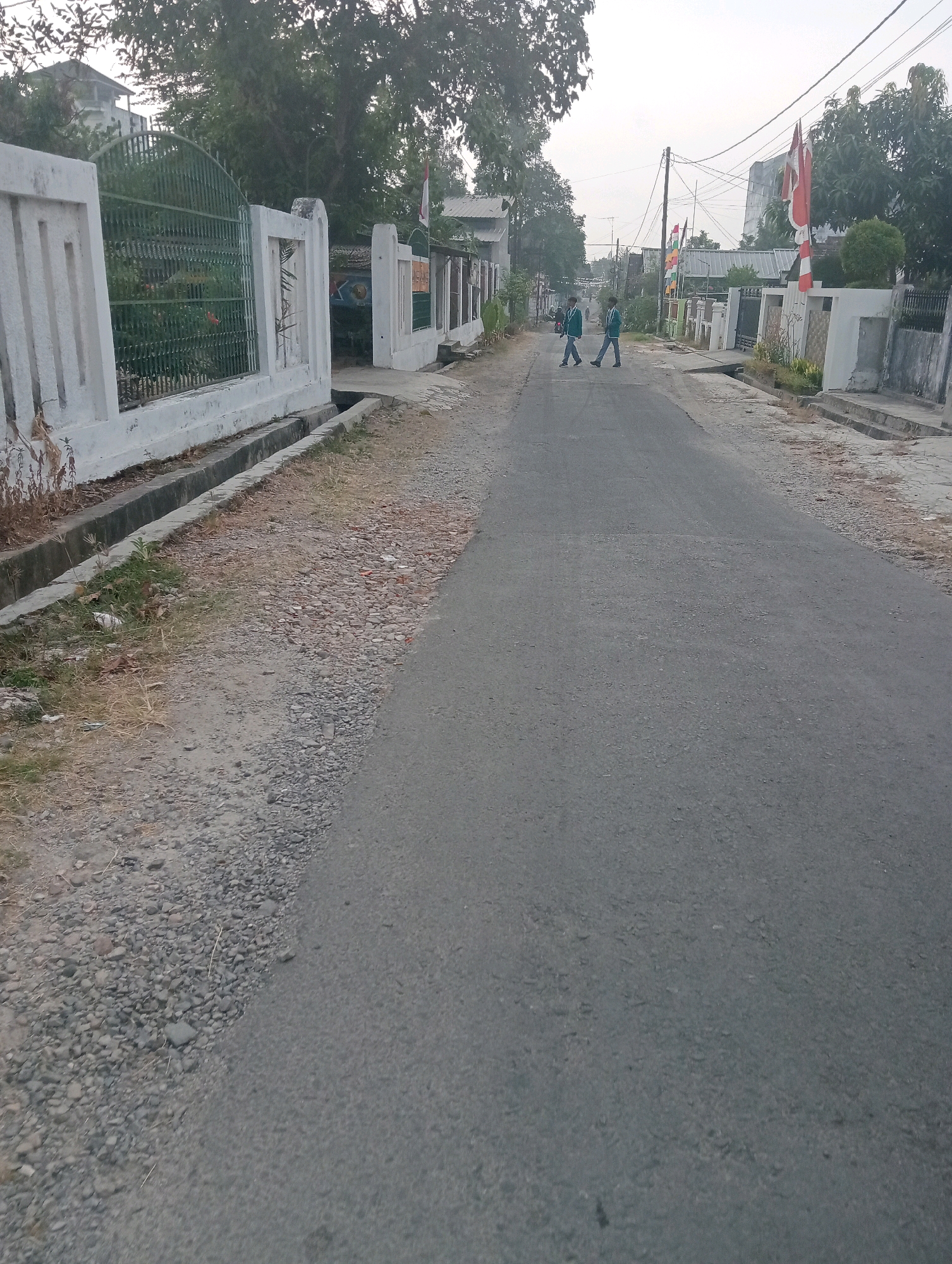 A straight, paved road runs through a residential area in Pringsewu, Indonesia. Two young boys, presumably students in uniform, are walking across the road from right to left. They are wearing dark blue and green jackets and pants. The road is flanked by a concrete drainage ditch on the left and a white concrete fence and wall on the right. Behind the fence on the right are houses and trees, with several Indonesian flags visible. On the left side of the road, there is a green metal gate leading to a property with trees and foliage. The sky is overcast.