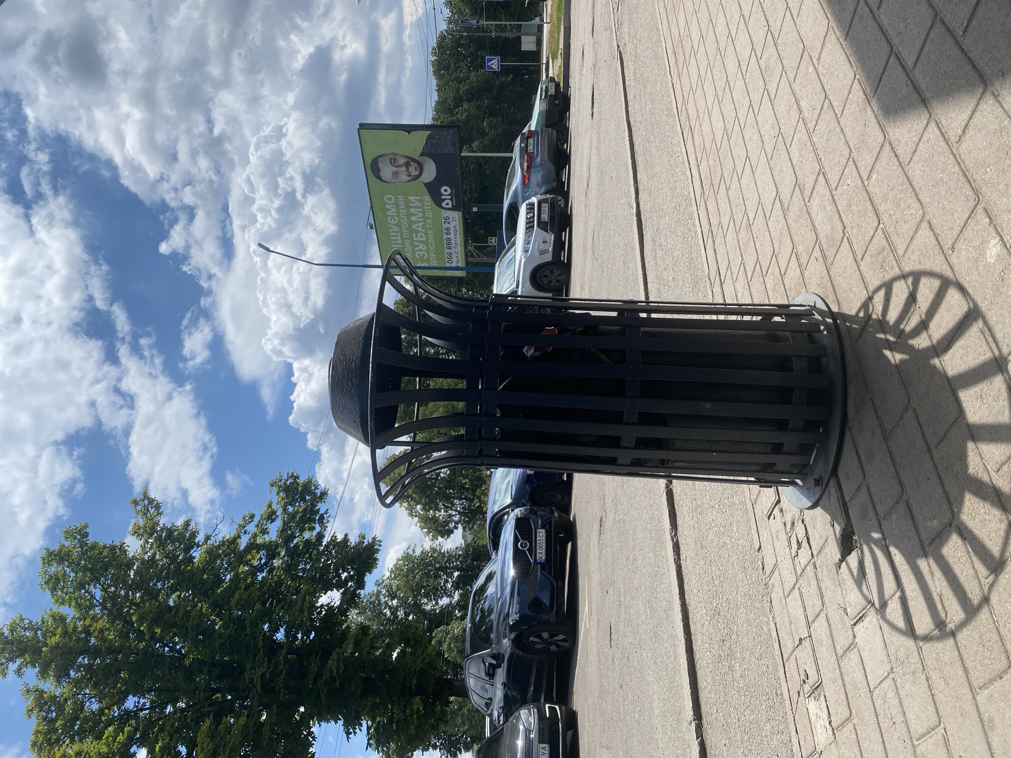 The image depicts an outdoor scene in Poltava, Ukraine, likely on a sunny day with scattered clouds. In the foreground, a large black metal trash receptacle stands on a paved sidewalk. The receptacle has a decorative, openwork design with vertical slats and a domed lid. Its shadow is cast on the patterned sidewalk. Behind the trash can, several cars are parked on an asphalt surface. One car, a dark blue Volvo SUV, is particularly visible, with a Ukrainian license plate reading "KA 0055 CT." Further in the background, a billboard advertising a dental clinic, "DIO DENTAL CLINIC," is displayed. The billboard features a man's portrait and Ukrainian text that translates to "We treat all dental problems for adults and children." A street lamp is visible near the billboard. Trees line the background, and distant buildings and a pedestrian crossing signal can be seen. The overall environment suggests an urban setting, possibly near a commercial area or parking lot. No people are actively engaged in any discernible actions in the frame.