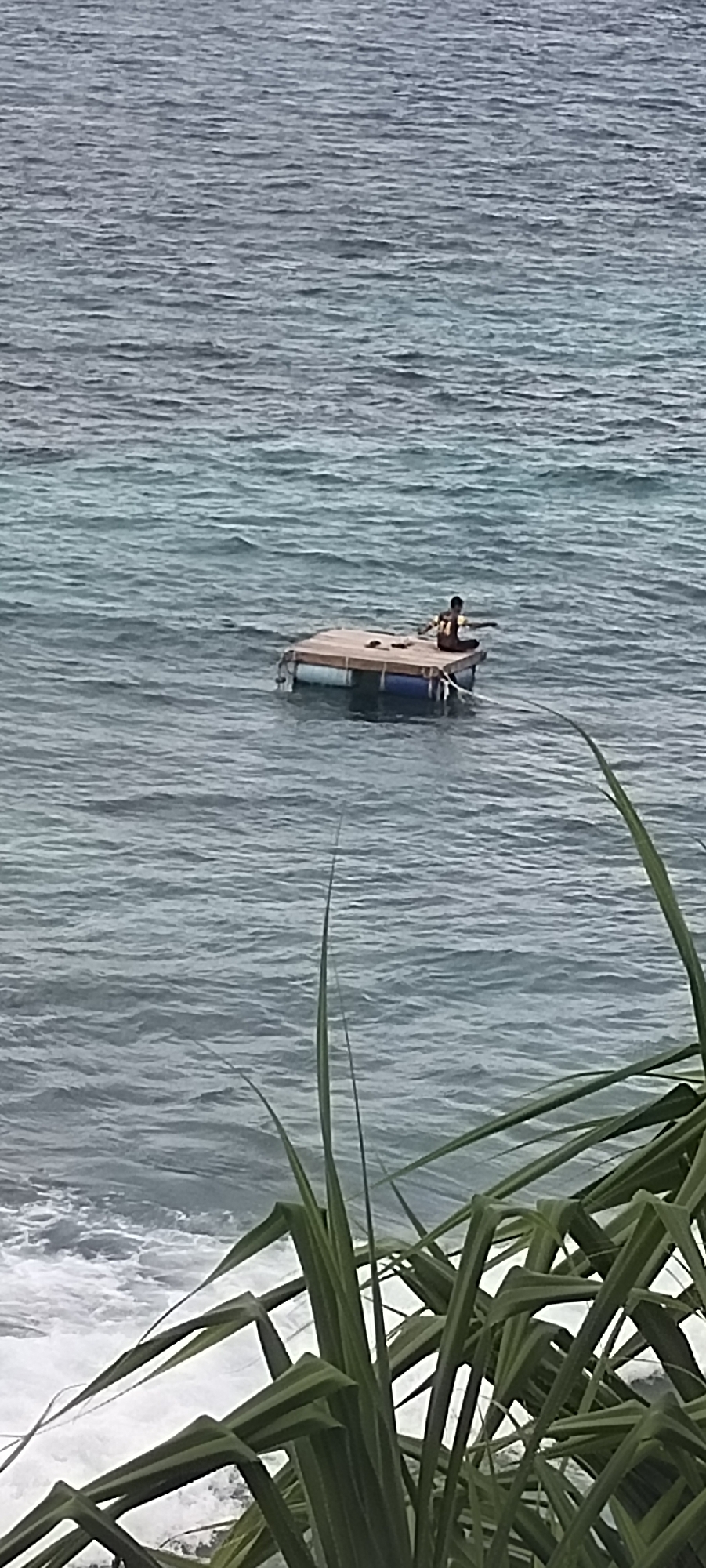 A lone individual sits on a makeshift raft in the choppy, grey waters of the ocean. The raft appears to be constructed from wooden planks supported by blue and white barrels. The individual, wearing a patterned shirt, is seated cross-legged in the center of the raft, facing away from the viewer and towards the right side of the frame. In the foreground, out-of-focus green foliage frames the scene, suggesting the vantage point is from land. The ocean is vast and appears to be somewhat rough, with visible waves. The overall impression is one of solitude and perhaps precariousness. This scene could be captured along the coastline of Sabang, Indonesia, known for its scenic beaches and maritime activities.