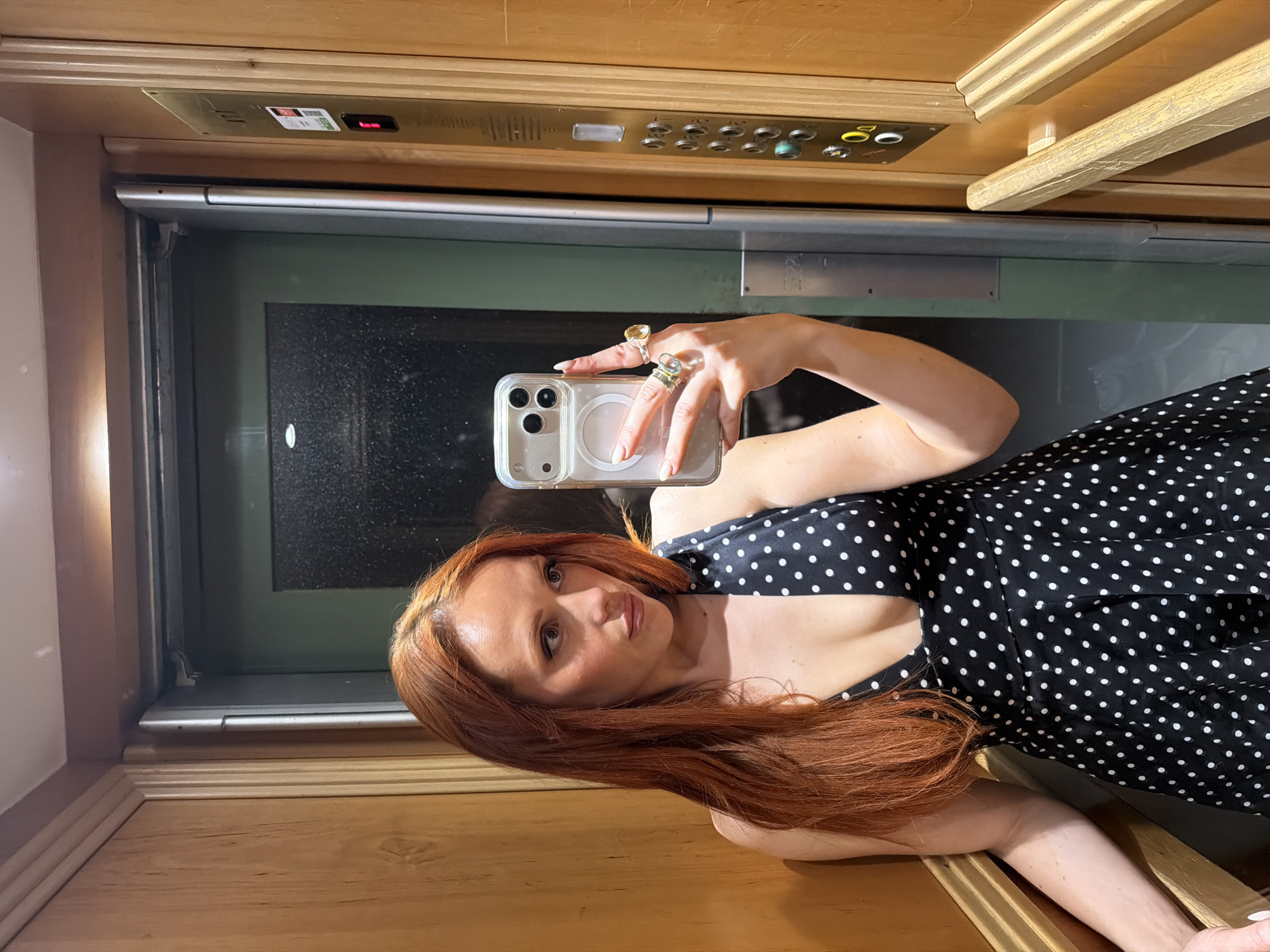 A woman with long, red hair is taking a selfie in what appears to be an elevator. She is holding a smartphone in her right hand, with her fingers adorned with rings. The phone is positioned to capture her face and upper body. She is wearing a black and white polka dot dress. Behind her, the elevator's interior is visible, including the buttons panel above and the door. The interior of the elevator is wooden, with a light grain. The scene suggests a modern, urban environment. There is no evidence to suggest the location is Paris, France.