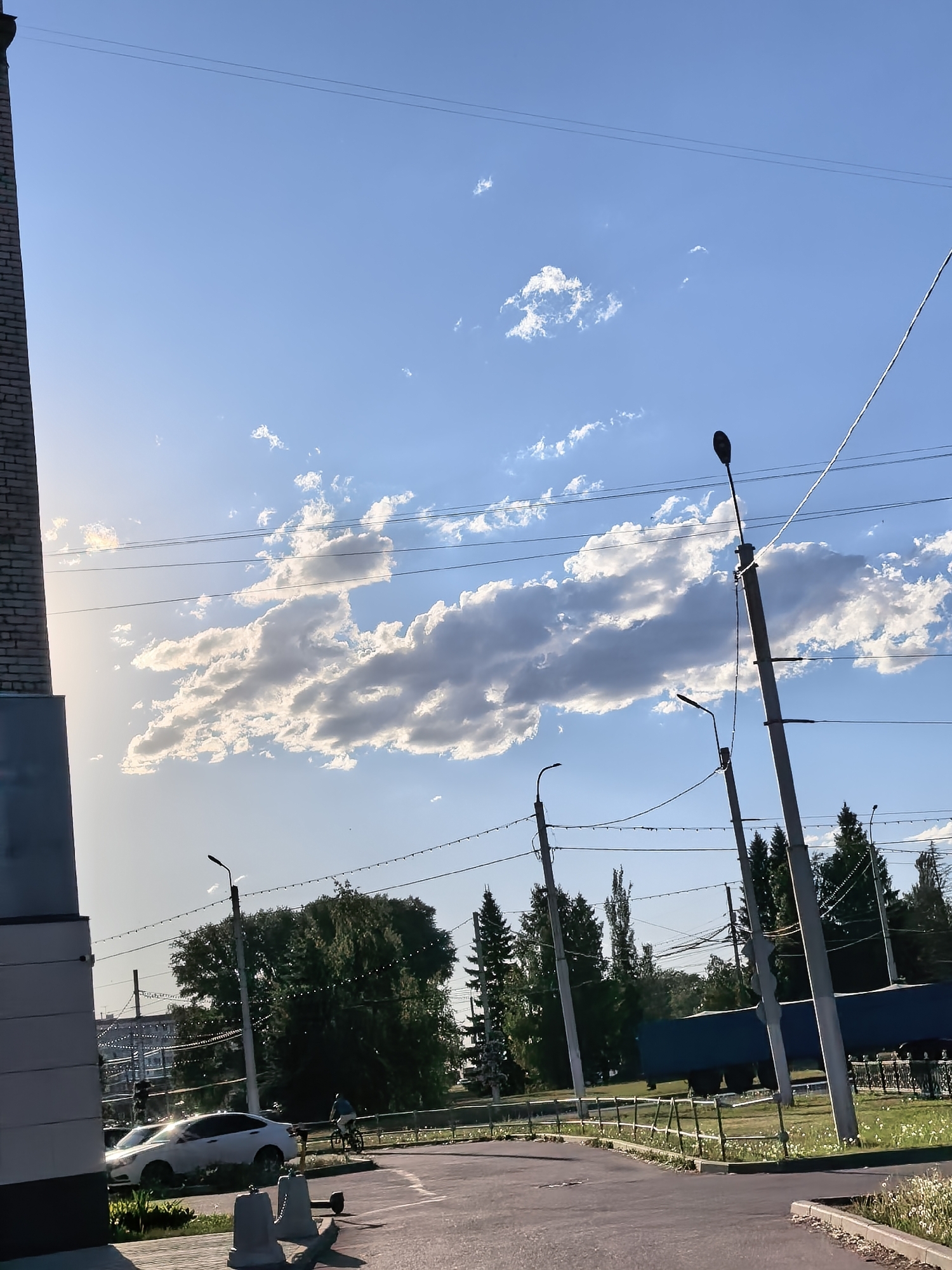 The image shows an urban scene in Sterlitamak, Russia. A white car is parked near a sidewalk, with a person riding a bicycle in the background. Overhead, power lines stretch across a blue sky with scattered clouds. Streetlights are visible along the road. Evergreen trees line the area behind the road. A grey and black brick building is partially visible in the left portion of the scene. The road surface appears to be paved.