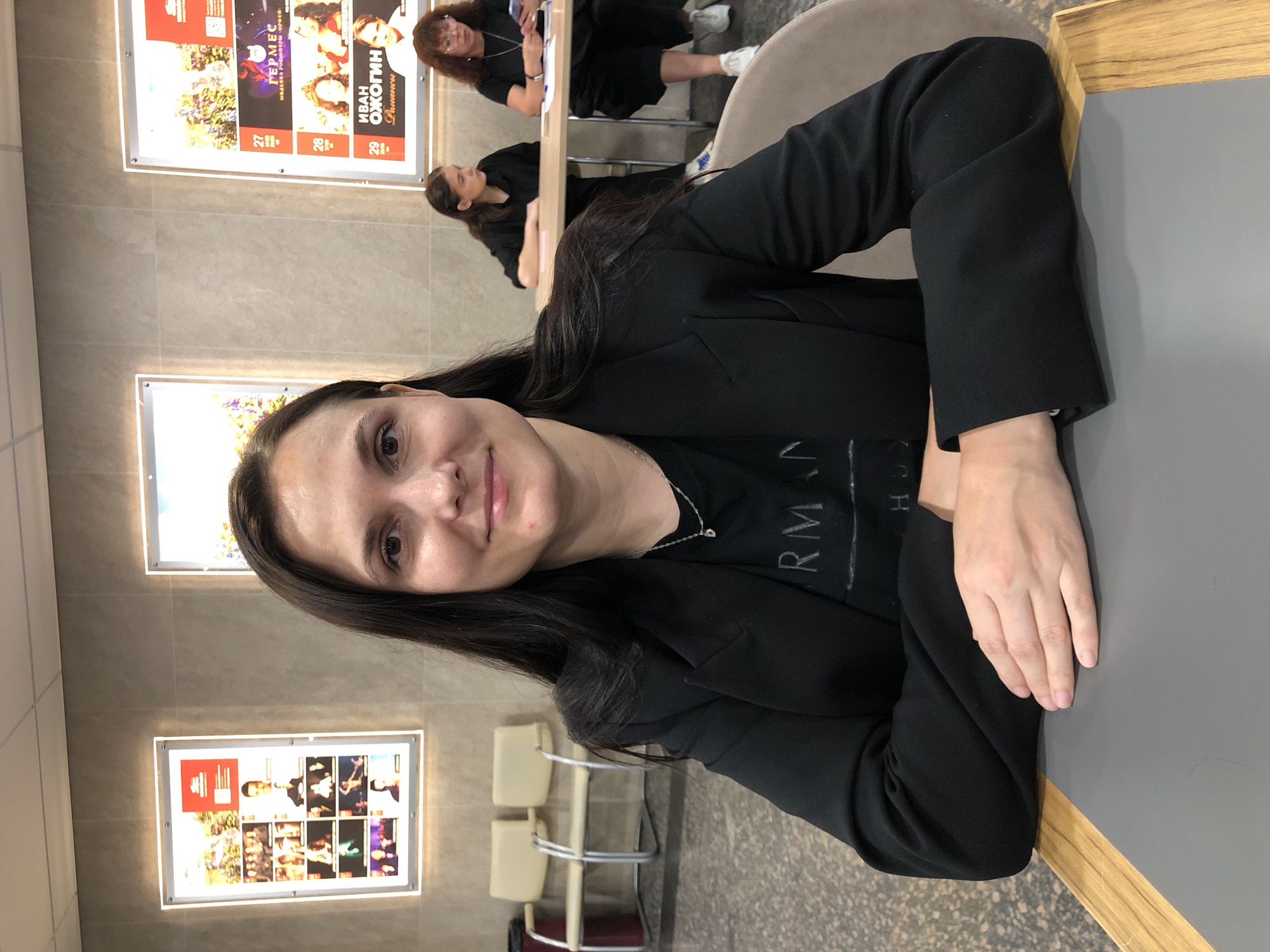 A medium shot shows a woman in a black blazer and t-shirt sitting at a grey table in what appears to be a public building, possibly a theater or exhibition hall, in Moscow, Russia. She has long, dark hair and is looking at the camera with a slight smile. Her hands are crossed in front of her on the table. In the background, two posters are visible on a wall; one features a collage of images and text, possibly an event advertisement, and the other displays an advertisement for an event with a photograph of a man. Two other women are seated at a table in the background, one looking at her phone and the other looking away from the camera. Two empty chairs and a trash can are visible to the left of the frame. The floor is speckled.