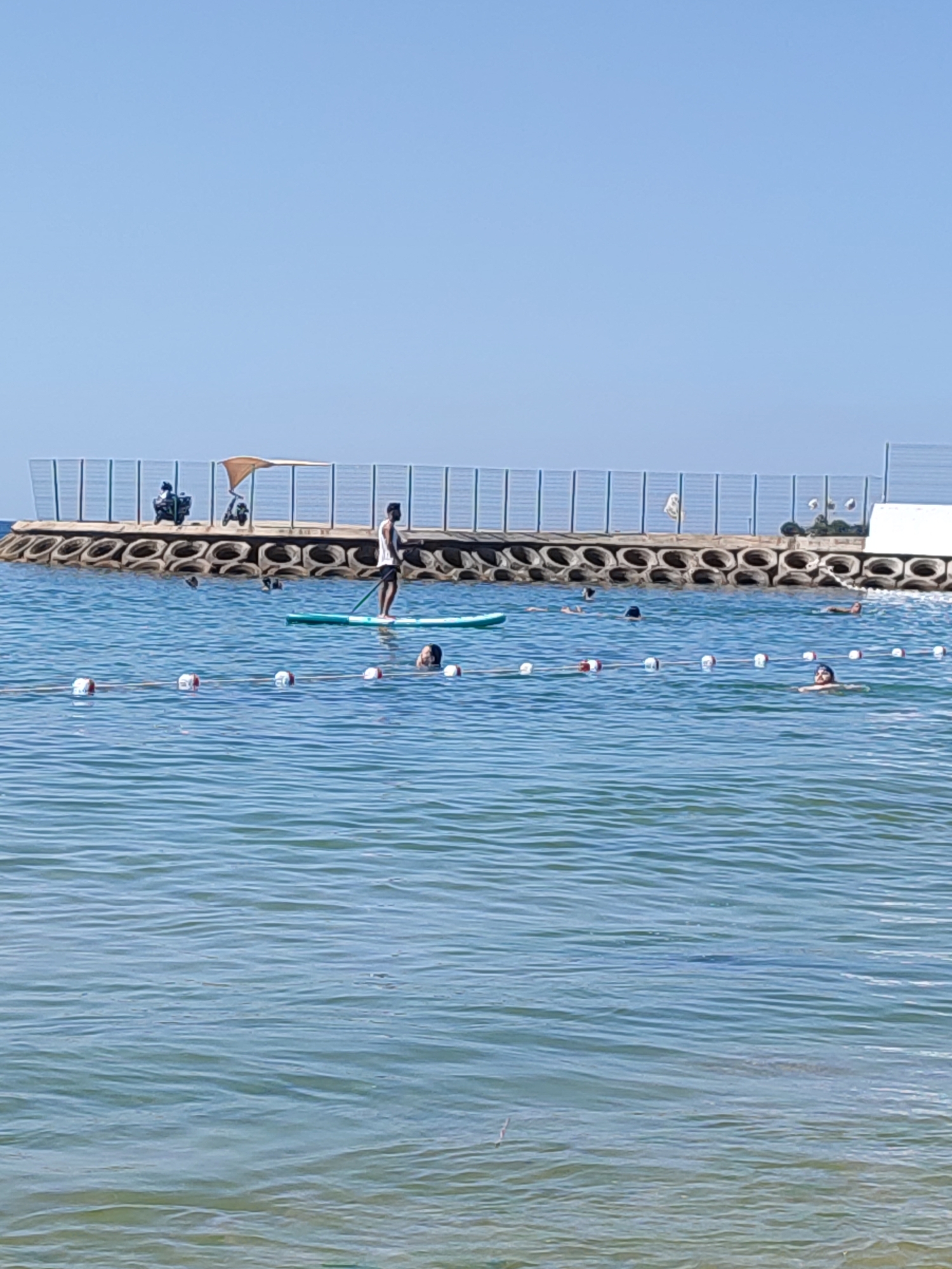 The image depicts a sunny day at a beach, likely in Kadıköy, Turkey, characterized by clear blue skies and calm sea water. In the foreground, the water is a bright turquoise, with subtle ripples indicating gentle movement. A rope with red and white buoys demarcates a swimming area.
Further back, several individuals are visible in the water. One person is stand-up paddleboarding on a green board, wearing dark shorts and a white tank top. Another person, with dark hair, is partially submerged in the water, looking towards the shore. Several other swimmers are scattered throughout the designated area, some with their heads above water, others less visible.
In the background, a breakwater extends into the sea, constructed from concrete blocks and what appear to be tires arranged in a circular pattern. Along the breakwater, a small beige awning shades a resting area, and two scooters are parked. A metal fence runs along the top of the breakwater, separating it from a white structure on the far right, possibly a building or a changing room. The overall scene conveys a relaxed atmosphere of recreational activity at the seaside.
