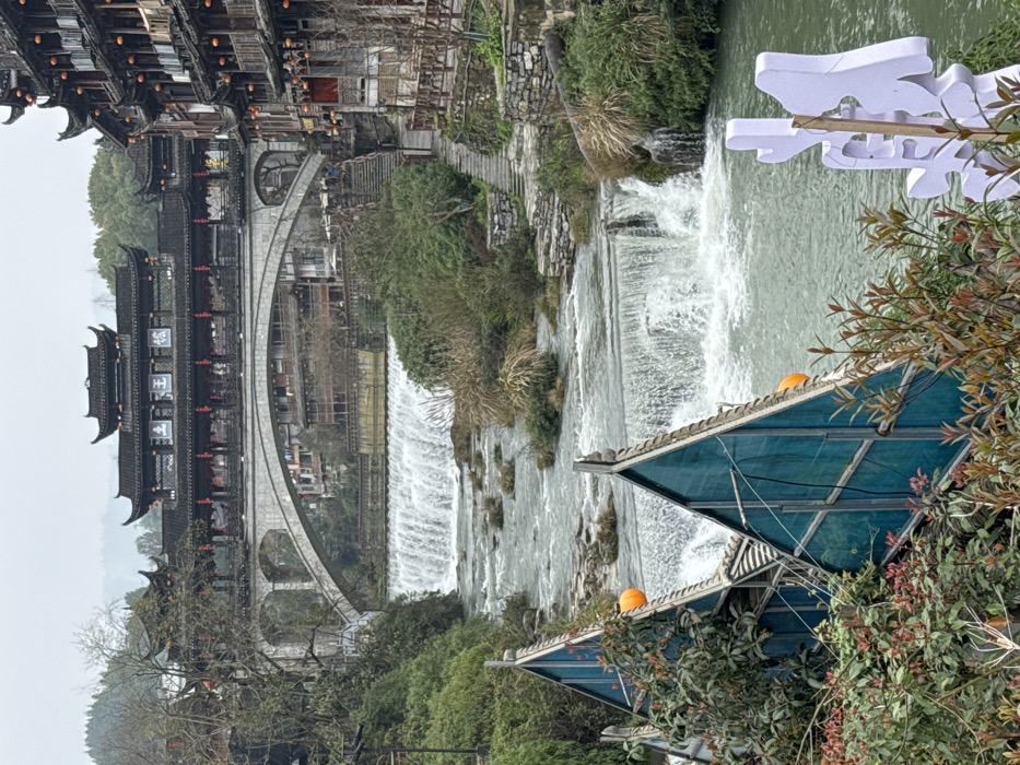 The image depicts a panoramic view of Furong Ancient Town, China, under an overcast sky. A river with light green water flows through the center of the frame, featuring multiple natural and man-made cascades forming distinct waterfalls. Small islands of green and brown vegetation are scattered within the riverbed.
In the immediate foreground, dense green and reddish-brown foliage partially obscures two structures with steep, triangular teal-colored panel roofs and dark frames, suggesting small shelters or boathouses. Orange spherical objects, possibly lanterns, are visible near these structures. A white, stylized Chinese character sign is partially visible on the bottom right.
The right bank of the river features stone steps leading uphill, flanked by stone walls and wooden railings. Adjacent to these steps are multi-story traditional Chinese wooden buildings with dark tiled roofs, intricate architectural details, and numerous red lanterns hanging from their facades. The left bank is densely vegetated with trees and shrubs.
Dominating the midground and background is a large stone arch bridge with at least three visible arches, spanning the river. This bridge supports an extensive, ornate traditional Chinese building, likely a covered bridge or pavilion, characterized by multiple dark-tiled roofs, red pillars, and red lanterns. White signs with Chinese characters are visible on its facade. Beyond the bridge and the town's structures, forested hills rise under the gray sky. No individuals or specific actions are visible within the scene.