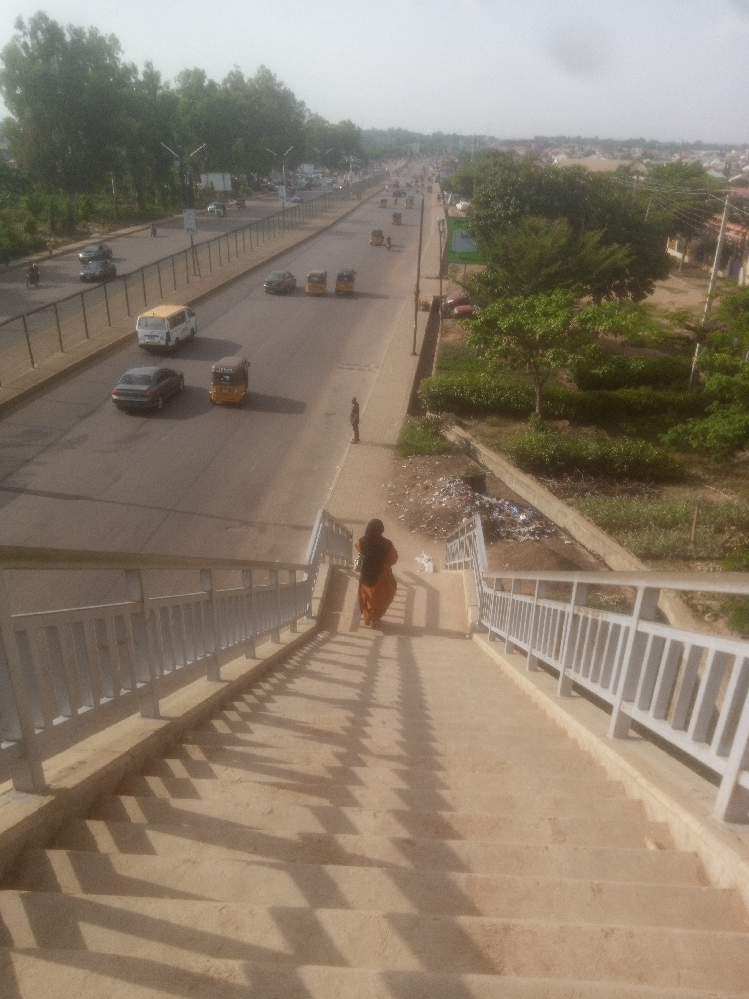 The image shows an elevated perspective looking down a concrete staircase with metal railings, likely part of a pedestrian overpass in Unguwan Dosa, Nigeria. A woman wearing an orange dress is descending the stairs, casting a shadow on the steps. Below the staircase, a multi-lane road runs parallel to lush green trees and vegetation. Several vehicles are visible on the road, including a white van, a dark-colored sedan, and two yellow tricycles. In the background, buildings and more trees suggest an urban environment. To the right of the road, there is a grassy area with a pile of garbage. The sky is overcast with a light haze.