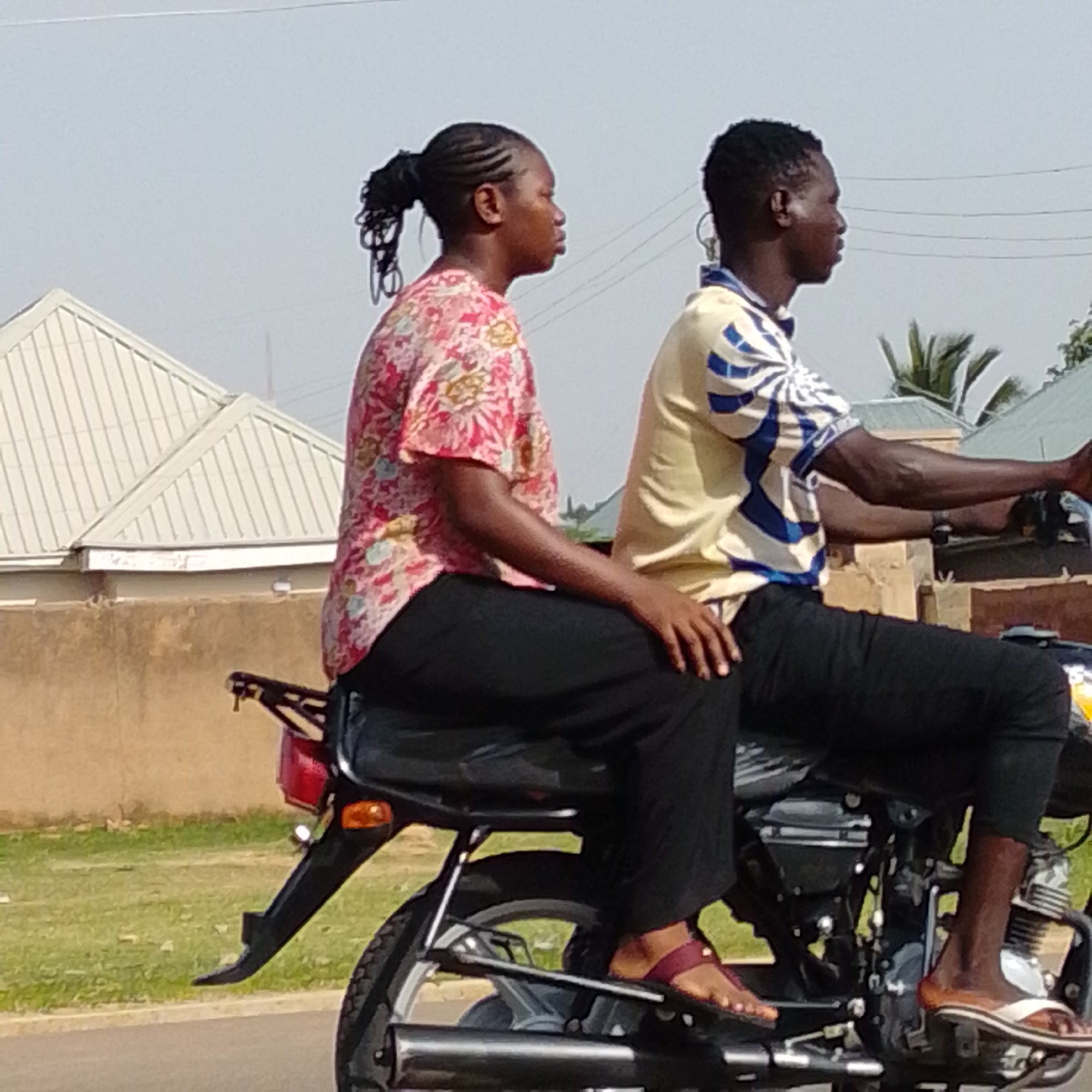 This is a medium shot image taken on a sunny day, depicting two people riding a motorcycle. The woman in the foreground, seated behind the driver, is wearing a vibrant, floral-patterned short-sleeved shirt and black trousers. Her hair is styled in braids pulled into a ponytail. The man in front, operating the motorcycle, is wearing a yellow and blue striped short-sleeved shirt and black trousers. The motorcycle itself is dark-colored. In the background, there are buildings with corrugated metal roofs, a brown wall, and green grass. The sky is clear and light blue. The scene appears to be in an urban or semi-urban environment, possibly a street in Gombe, Nigeria, given the context. The image captures a common mode of transportation in such locations.