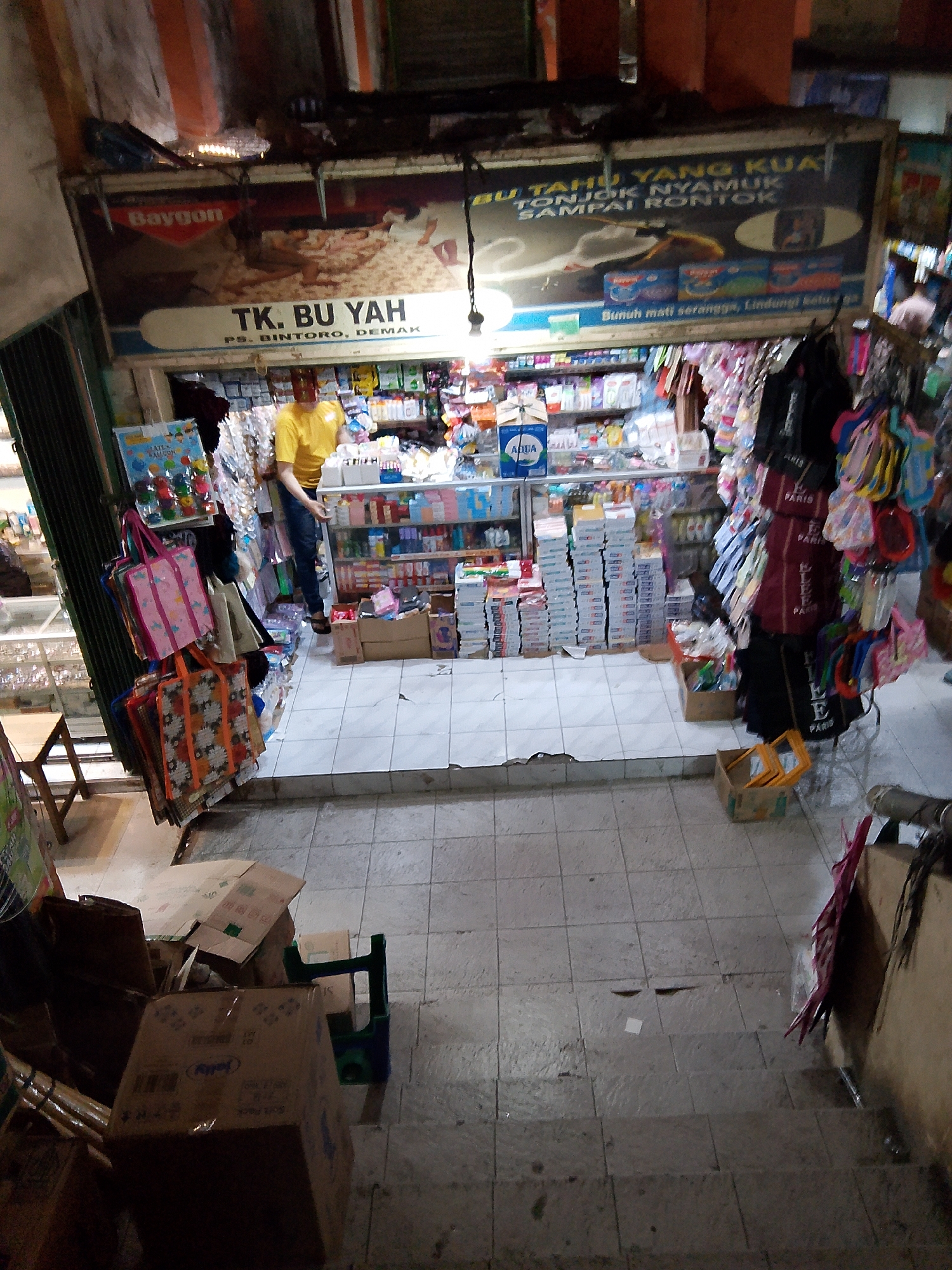 The image captures a street-level view of a small shop in Bintoro, Demak, Indonesia. The shop appears to be a convenience store or a general store, selling a variety of household items. The storefront is open, revealing shelves stocked with packaged goods, bottles, and other products. A man wearing a yellow t-shirt and blue jeans stands inside the shop, seemingly attending to business. In front of the shop, colorful tote bags and other merchandise are displayed on racks. The floor is tiled, and there are some cardboard boxes and a green plastic stool visible. The shop's signage, partially visible, indicates "TK. BU YAH" and "PS. BINTORO, DEMAK." A large advertisement poster hangs above the shop's entrance, featuring text in Indonesian. The overall impression is that of a bustling local market or retail establishment.