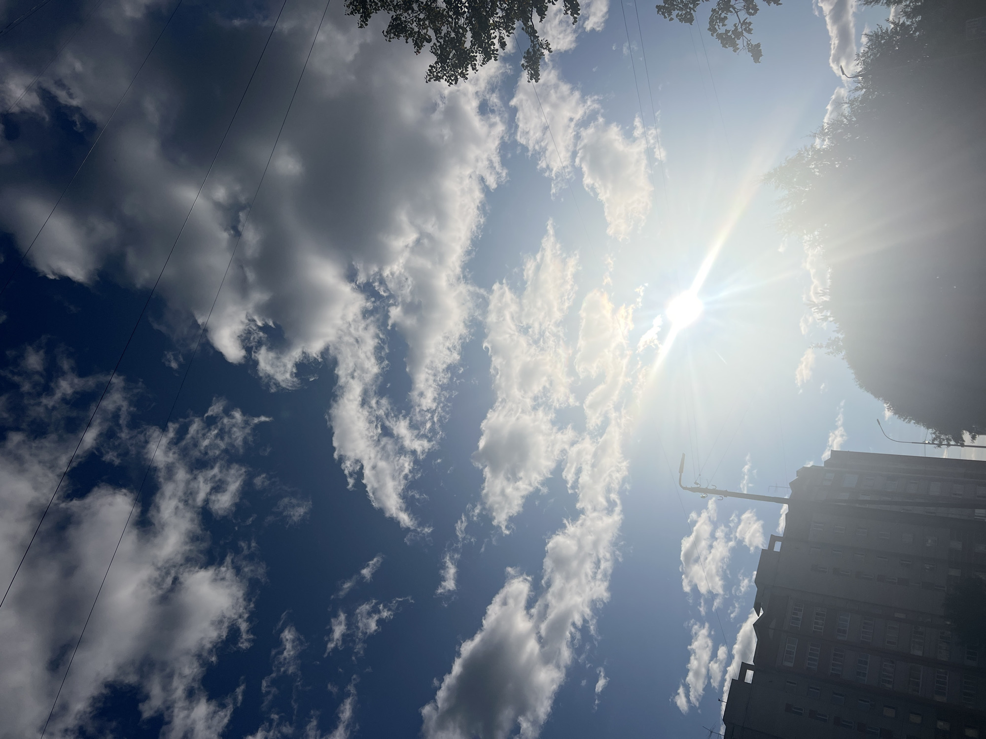 The image depicts a bright, sunny day in an urban environment, possibly in the city of Unknown, Unknown. The sky is a vibrant blue, dotted with scattered white and gray clouds. The sun is shining intensely, casting a bright glare and sunbeams across the scene. In the foreground on the left, a multi-story apartment building stands tall, its facade detailed with numerous windows. To the right and lower portion of the image, the silhouette of trees is visible, partially obscured by the sun's glare. Power lines stretch across the sky, intersecting with the clouds. A street lamp is also visible near the apartment building. The overall impression is of a typical day in a city, with a mix of residential buildings, natural elements, and infrastructure under a bright sky.