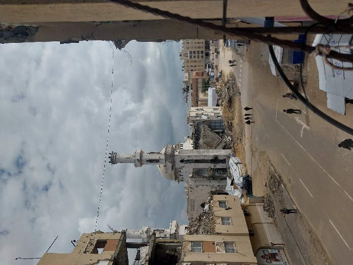 The image depicts a street scene in Khan Yunis, Palestinian Territory, characterized by widespread structural damage under an overcast sky. At the center, the Great Mosque of Khan Yunis (Al-Kabir Mosque) is heavily damaged, its white minaret and dome partially intact, but large sections of its main structure have collapsed into piles of rubble around its base. Multi-story buildings to the left of the mosque are severely damaged, with upper floors partially destroyed, exposing interiors and twisted rebar. An asphalt road, with visible white lane markings and a turn arrow, traverses the foreground and mid-ground, bordered by dirt and debris.
Approximately 12-15 individuals are present on the street and sidewalks. A group of four people walks together across the main intersection near the mosque. Three individuals stand on the street in the mid-ground, while another person stands on the left sidewalk. An individual walks in the immediate foreground, partially obscured, and other figures are visible in the distant background. A dark-colored car is parked amidst rubble near the mosque. On the lower section of the damaged building on the left, graffiti and a red square graphic are visible. Small, makeshift stalls with corrugated metal roofs are present on the right side of the foreground. Cables stretch overhead across the scene between damaged buildings. A metal railing frames the right edge of the image.