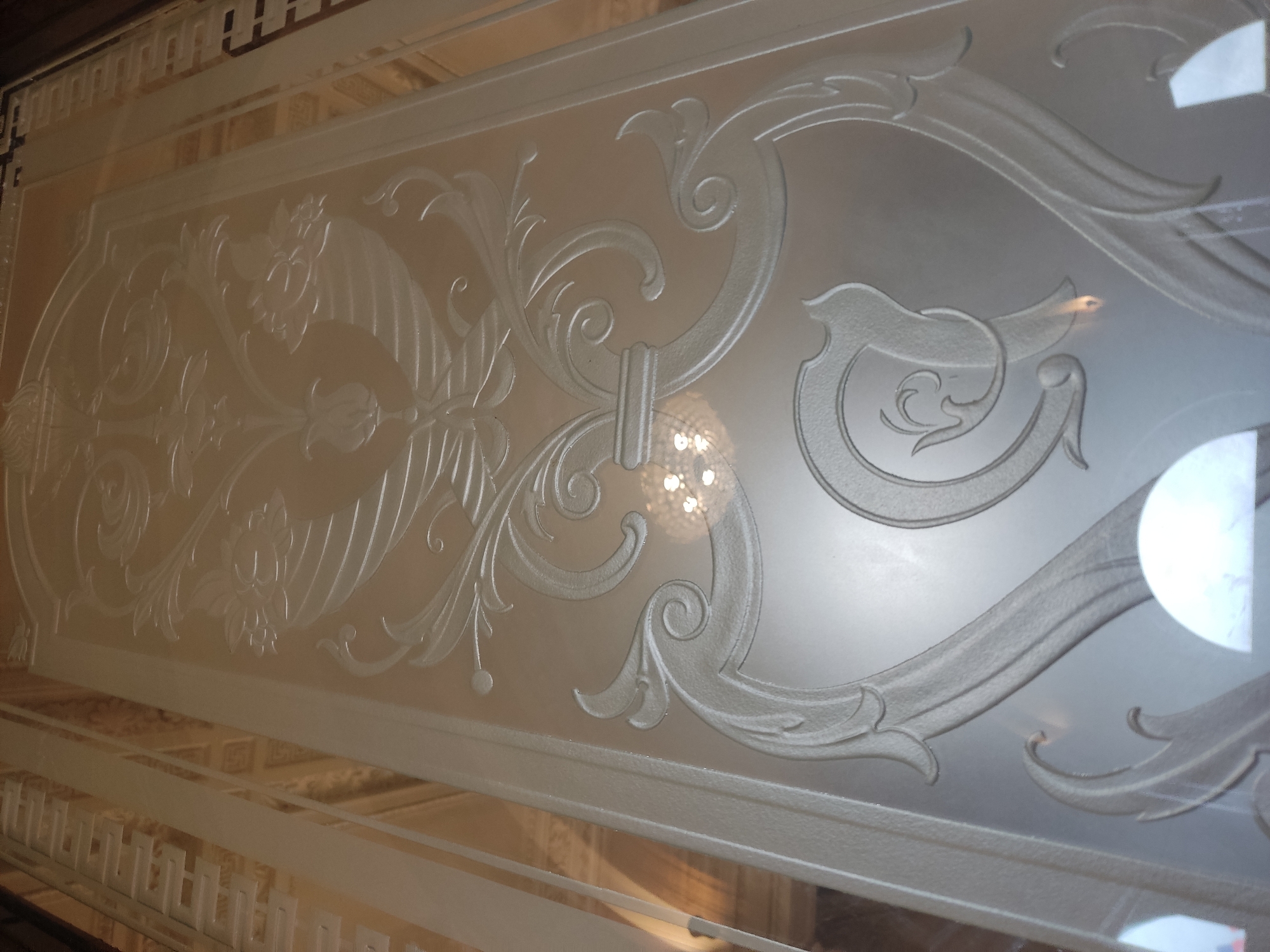 The image shows a close-up of a frosted glass panel with ornate floral and scrolling designs. The panel is likely part of an architectural feature, possibly a door or window, and is back-lit, revealing details of the etched or sandblasted design. The location is unknown.
