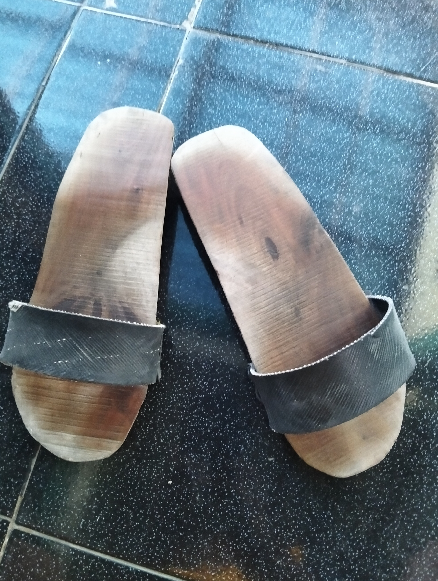 A pair of wooden clogs with black straps rests on a dark, tiled floor. The clogs are made of light-colored wood with visible grain patterns and appear worn. The black straps are textured and wrap around the front of each clog. The floor is composed of square tiles with a speckled pattern. This image was captured in Paciran, Indonesia.