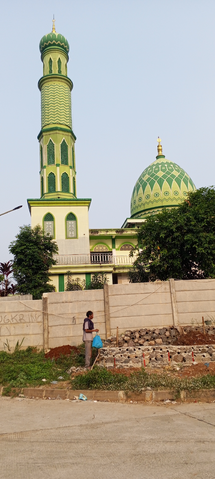 An exterior shot captures a mosque with a green, multi-tiered minaret and a green dome, both adorned with intricate geometric patterns. The building itself is cream-colored with green accents around the windows and balconies. In the foreground, a person stands near a concrete wall, holding a blue bag. The area behind the wall appears to be undergoing construction, with piles of rocks and exposed earth. Greenery, including trees and bushes, surrounds the mosque and the construction site. The scene is set against a clear, pale blue sky. The mosque is located in Depok, Indonesia.