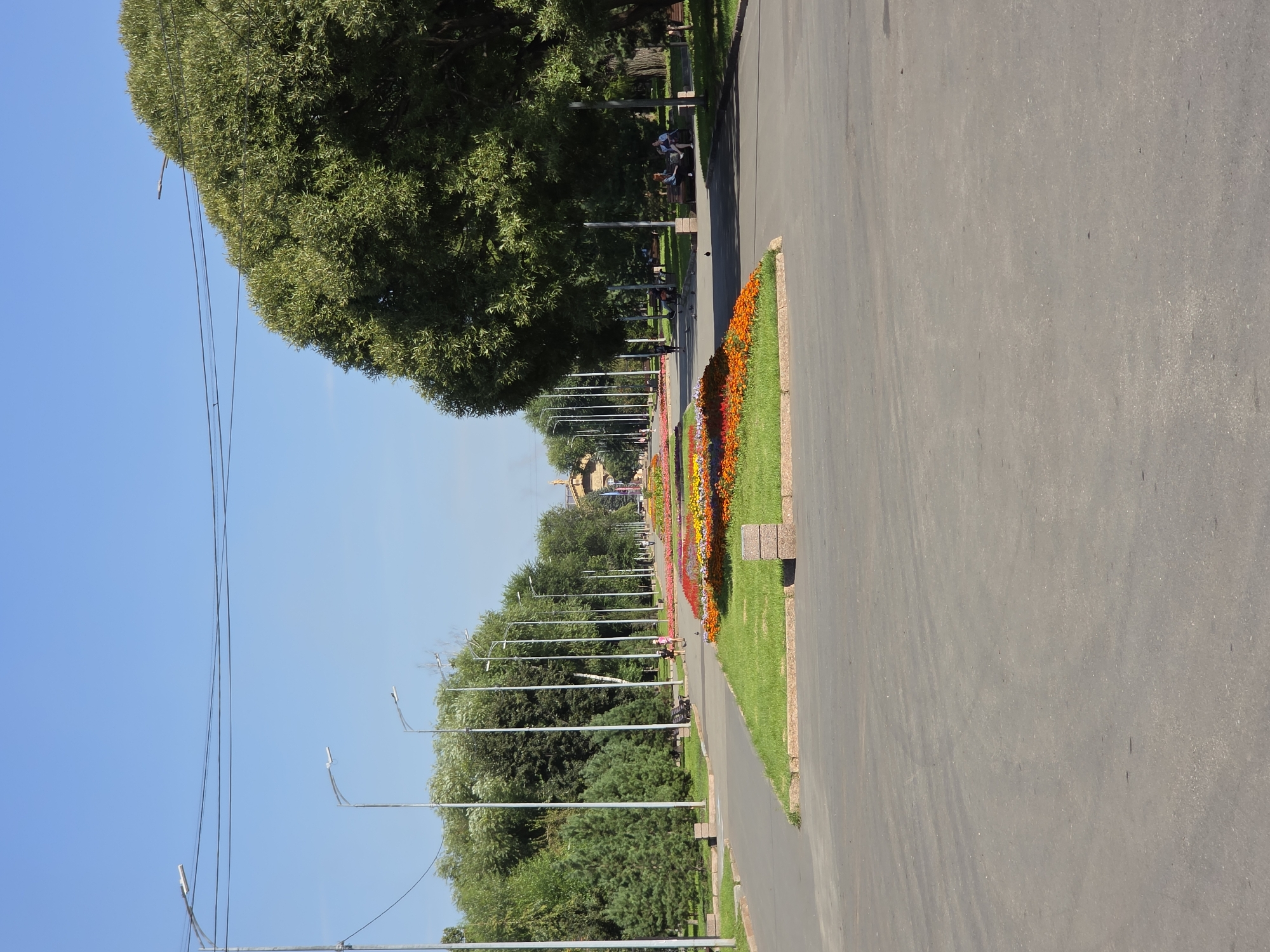 This image depicts an outdoor scene, likely a park or a wide pedestrian promenade, in Chelyabinsk, Russia. The foreground features a paved pathway and grass borders, with a central section containing a long, elaborate flowerbed filled with red, orange, and yellow flowers. Beyond this, the pathway continues, flanked by rows of streetlights and mature trees on both sides. In the middle ground, several people are visible walking and sitting on benches, enjoying the sunny day. The background shows more trees and a large, ornate building or monument in the distance. The sky is clear and blue.
