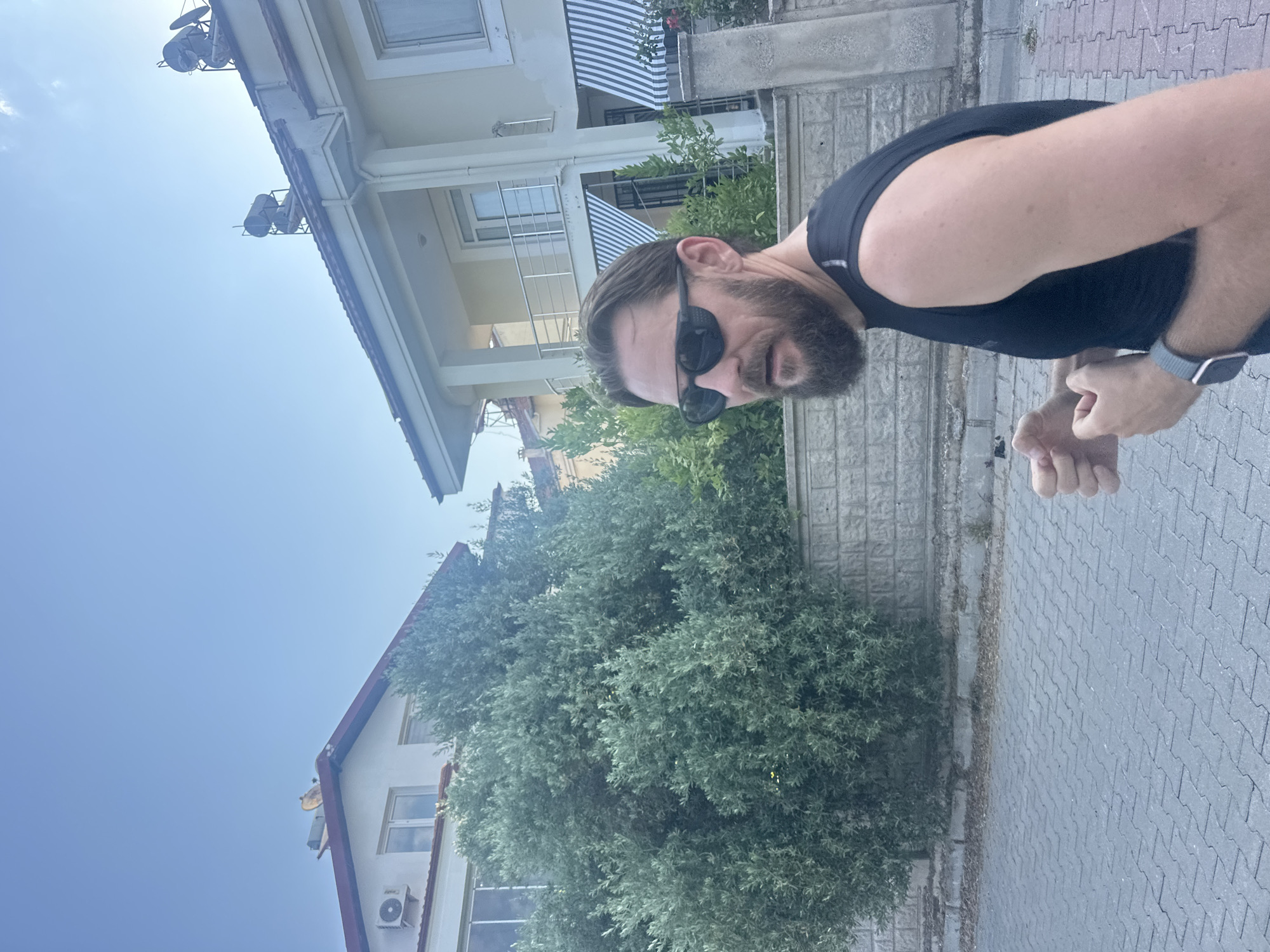In Fethiye, Turkey, a man with a beard and sunglasses is captured in a medium shot, facing right. He is wearing a black sleeveless shirt and a watch on his left wrist. The man is standing on a paved walkway, with a stone wall and lush greenery behind him. In the background, there are two houses with solar water heaters on their roofs. The sky is clear blue. The scene appears to be an outdoor setting in a residential area, possibly during the day given the clear sky and sunlight.
