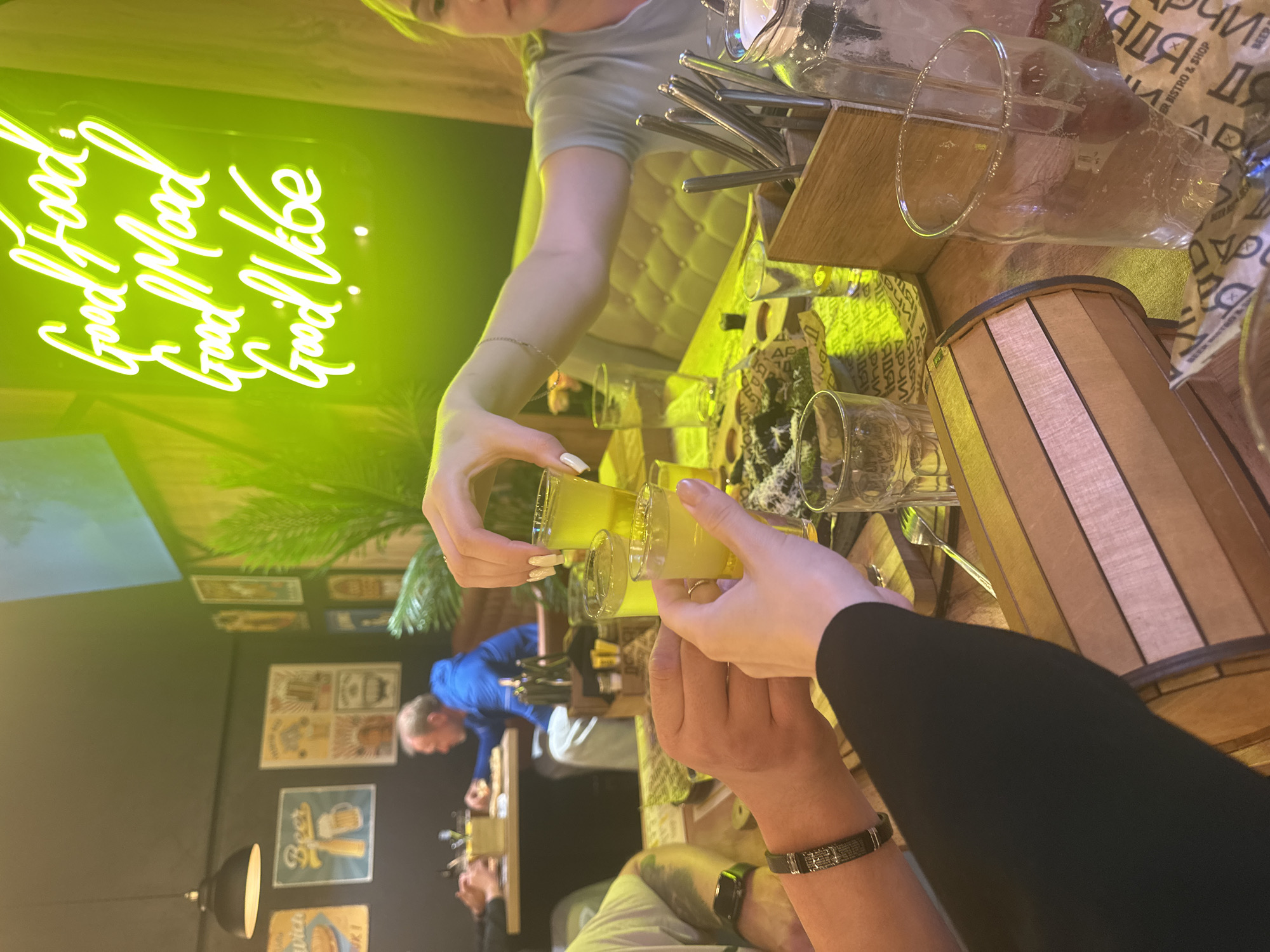 Here is a forensic description of the image:
**Scene:**
The image depicts an indoor gathering at a restaurant or bar, likely in Perm, Russia. This is suggested by the presence of the Cyrillic text "Пермь" on what appears to be a beer-related flyer in the background. The scene is brightly lit, with a warm, yellowish hue. The focal point is a group of people raising small glasses (shots) of a yellow-colored liquid, possibly a liqueur or cocktail, for a toast. A neon sign with the words "Good Food, Good Mood, Good Vibe" is visible on the left, casting a green glow.
**People:**
At least four individuals are partially visible:
1. A person in a light-colored top is pouring the yellow liquid into the shot glasses.
2. Two people in the foreground are holding up glasses for a toast, one wearing a bracelet.
3. Another person in the background is wearing a blue top.
**Objects:**
* Several shot glasses with a yellow liquid.
* A wooden caddy containing cutlery (forks and spoons) and possibly other utensils.
* A wooden container.
* Bottles and other items on a counter or table in the background.
* Various beer-related posters, possibly advertising "Beer Bistro & Shop."
**Actions and Interactions:**
The primary action is the toast, as indicated by the raised shot glasses. The interaction between people suggests a celebratory or social occasion.
**Environment:**
The environment is a well-lit restaurant or bar with a mix of modern and rustic elements. The neon sign and posters suggest a casual and lively atmosphere.