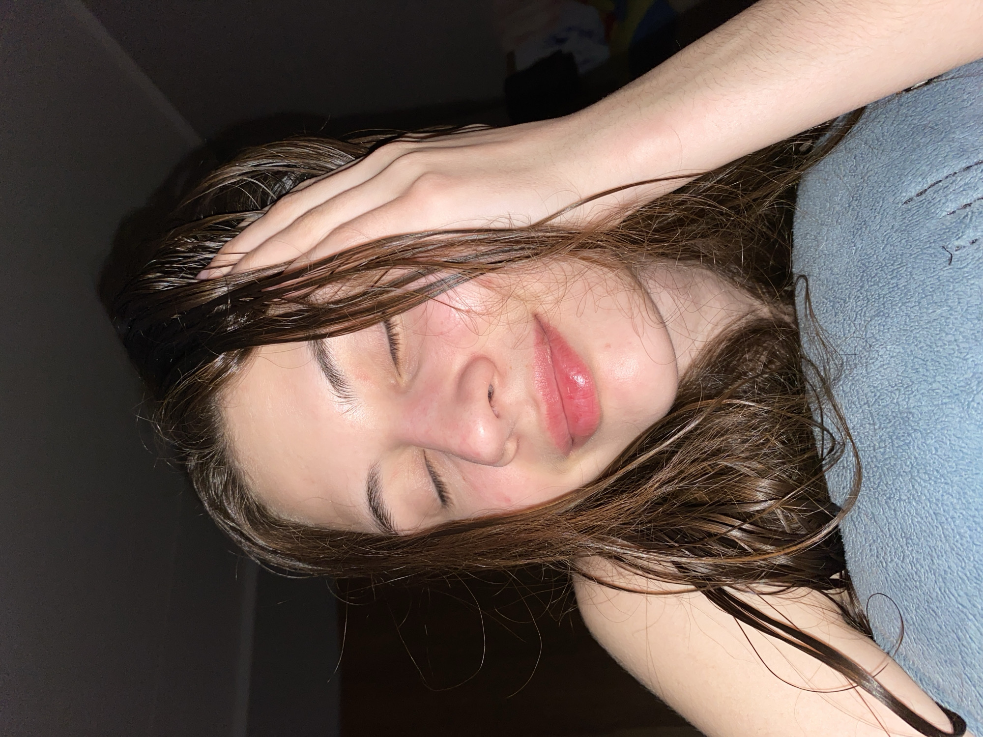 A close-up, dimly lit, portrait-style photograph shows a young person lying down, seemingly resting or asleep, in what appears to be an indoor setting in Votkinsk, Russia. The individual has closed eyes, suggesting relaxation or sleep. Their right hand is placed over their forehead, with fingers visible through their dark brown, wet-looking hair, which is spread across their face and shoulders. The hair appears to be recently washed, as it is glossy and clumped in strands.
The person's skin tone is fair, and there is a subtle pinkish flush visible on their cheeks and nose. Their lips are slightly parted, showing a natural pinkish hue, possibly with a hint of gloss. Visible facial features include well-defined eyebrows and the contours of their nose and lips.
The background is predominantly dark, indicating low light conditions, possibly night or a dark room. To the right side of the frame, a textured, light blue fabric, likely a pillow or blanket, is visible, partially obscured by the person's arm and hair. No specific location cues for Votkinsk, Russia are visually identifiable beyond the indoor, personal setting. There is no visible text in the image. The overall mood conveyed is one of quiet repose or exhaustion.