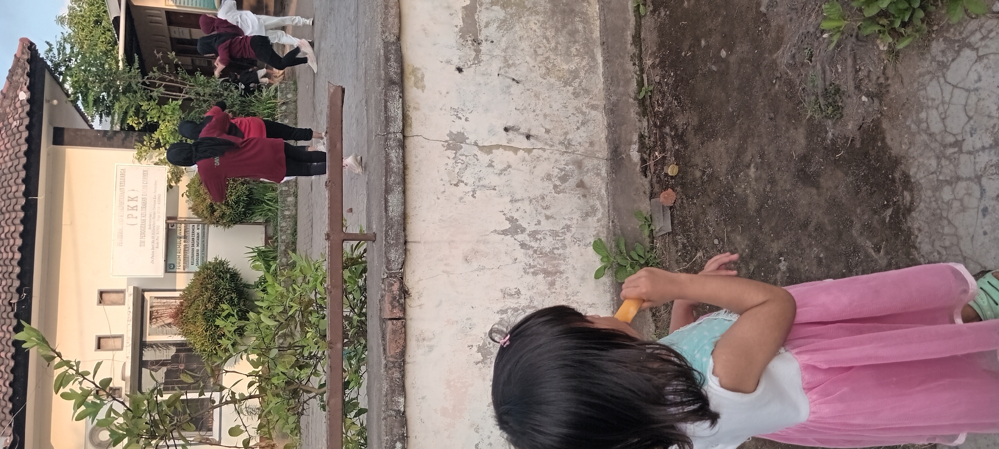 The image depicts an outdoor scene in Mataram, Indonesia, featuring a building with a sign for "TIM PENGERAK KELURAHAN DASAR CERMEN (PKK)" and a "FORUM REMBUG WARGA KAMPUNG SAPIR". Several individuals, predominantly women wearing dark headscarves and athletic attire, are engaged in what appears to be a group exercise or dance session in front of the building. The foreground is occupied by a young girl wearing a pink dress, facing away from the camera and eating a yellow object, possibly food or a toy. The environment consists of a paved walkway, a low concrete wall, and some vegetation. The building has a tiled roof and a light-colored facade. The lighting suggests it is daytime, possibly late afternoon, given the warm tones.