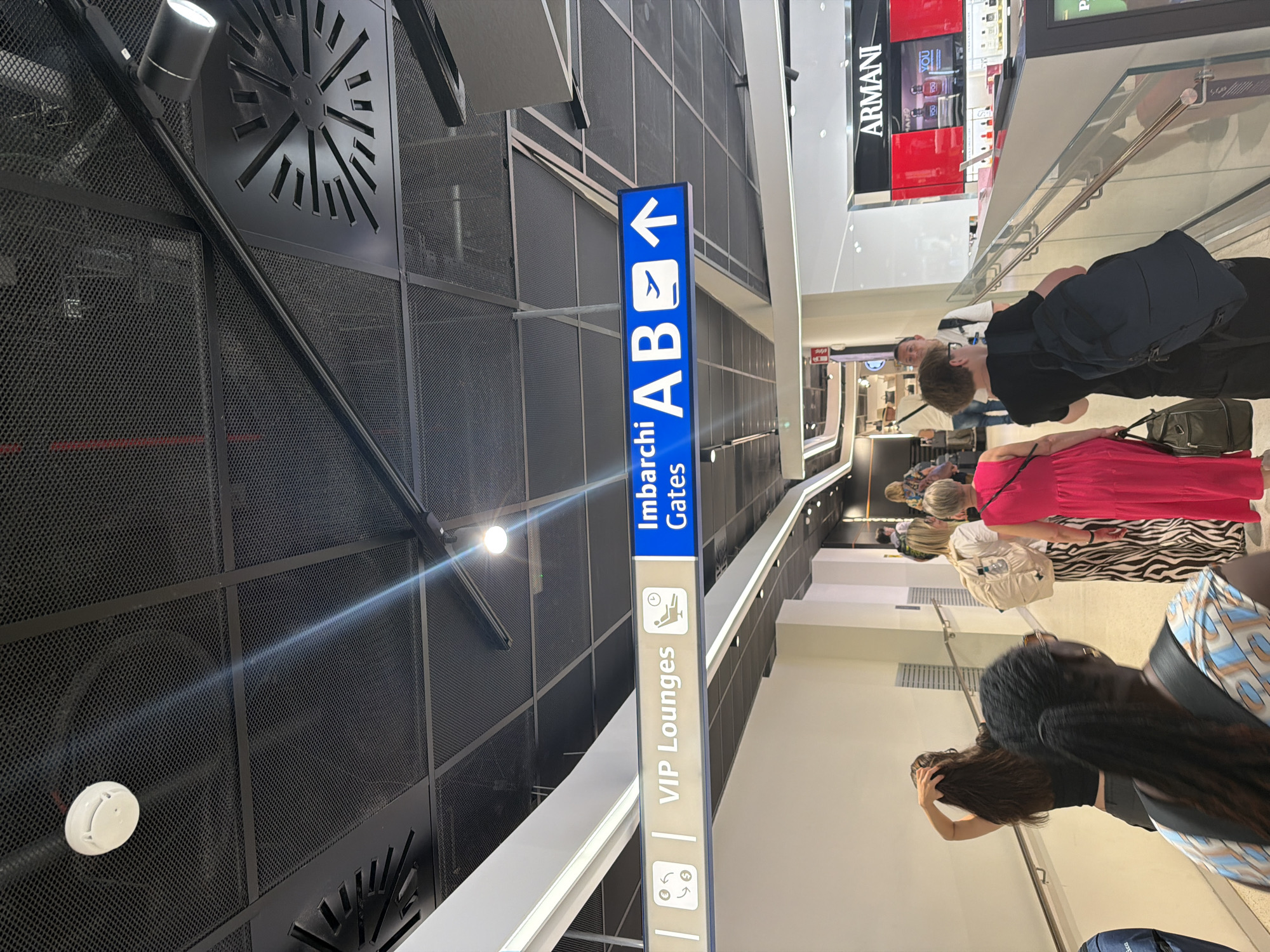 The image captures a view within an airport terminal, likely at Milan Linate Airport in Segrate, Italy, given the Armani store visible in the background. The scene depicts a group of people walking along a walkway. Overhead, there are directional signs. One sign, in blue with white lettering and icons, indicates "Imbarchi AB Gates" with an airplane and an upward arrow, suggesting departures for gates A and B. Another sign, in a lighter color, reads "VIP Lounges" and features icons representing currency exchange and a person relaxing. The ceiling is a dark, perforated metal grid, with track lighting and ventilation systems visible. Below, a railing separates the walkway from a lower level, where the Armani store is located. The people are dressed casually, some carrying backpacks and bags, indicating travel. The overall environment suggests a busy, modern airport concourse.