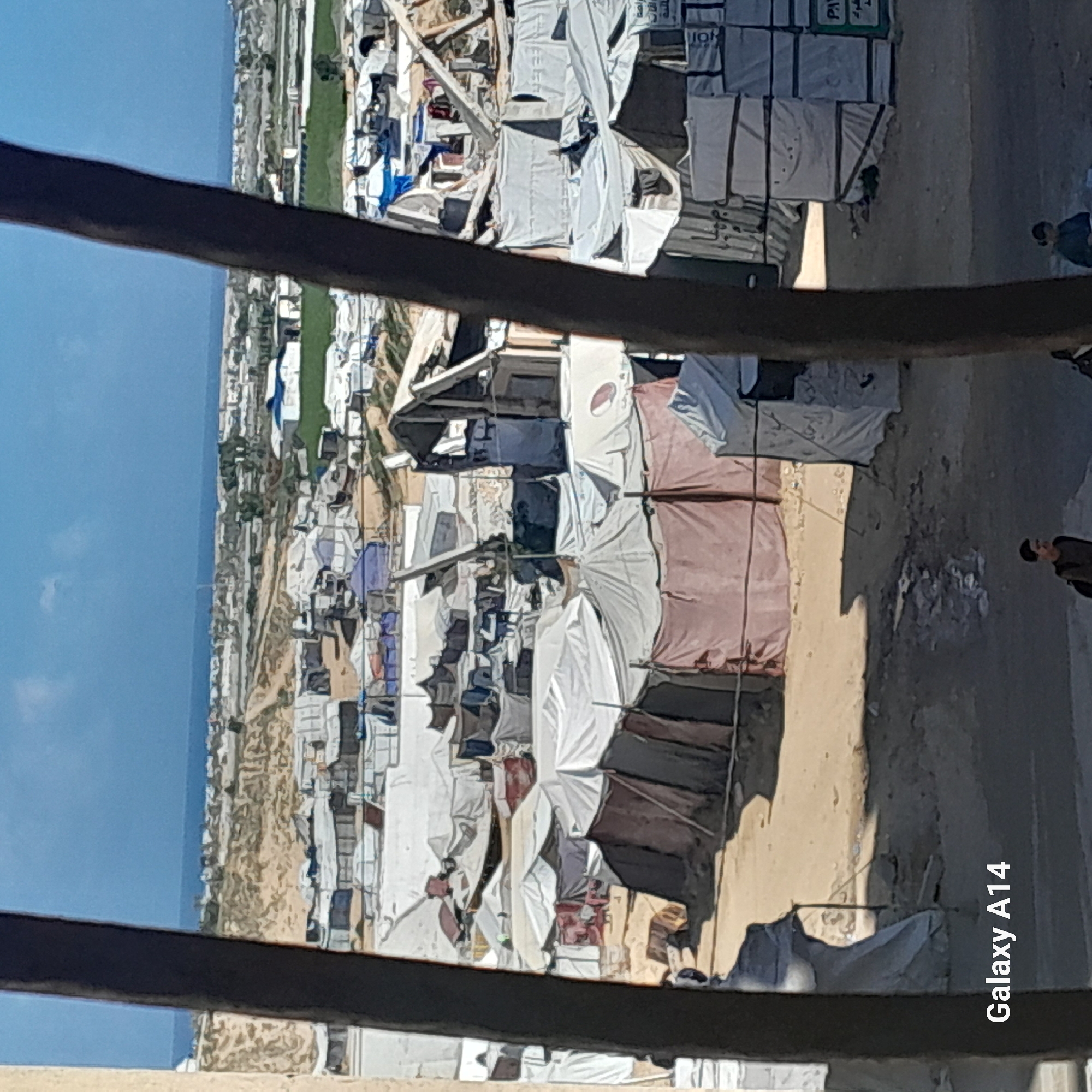 The image depicts a densely populated refugee camp in Khan Yunis, Palestinian Territory. Rows of makeshift tents, constructed from a variety of materials including tarps, cloth, and salvaged wood, dominate the landscape. Some tents have extensions or attachments, suggesting attempts to create more stable dwellings. A sandy, barren ground covers the camp. In the background, a body of water is visible near a strip of green vegetation. A steel structure is in the foreground, partially obscuring the view. Two people are visible on the far right. The overall impression is one of impoverishment and precarious living conditions. The lower right corner of the image has the text "Galaxy A14."
