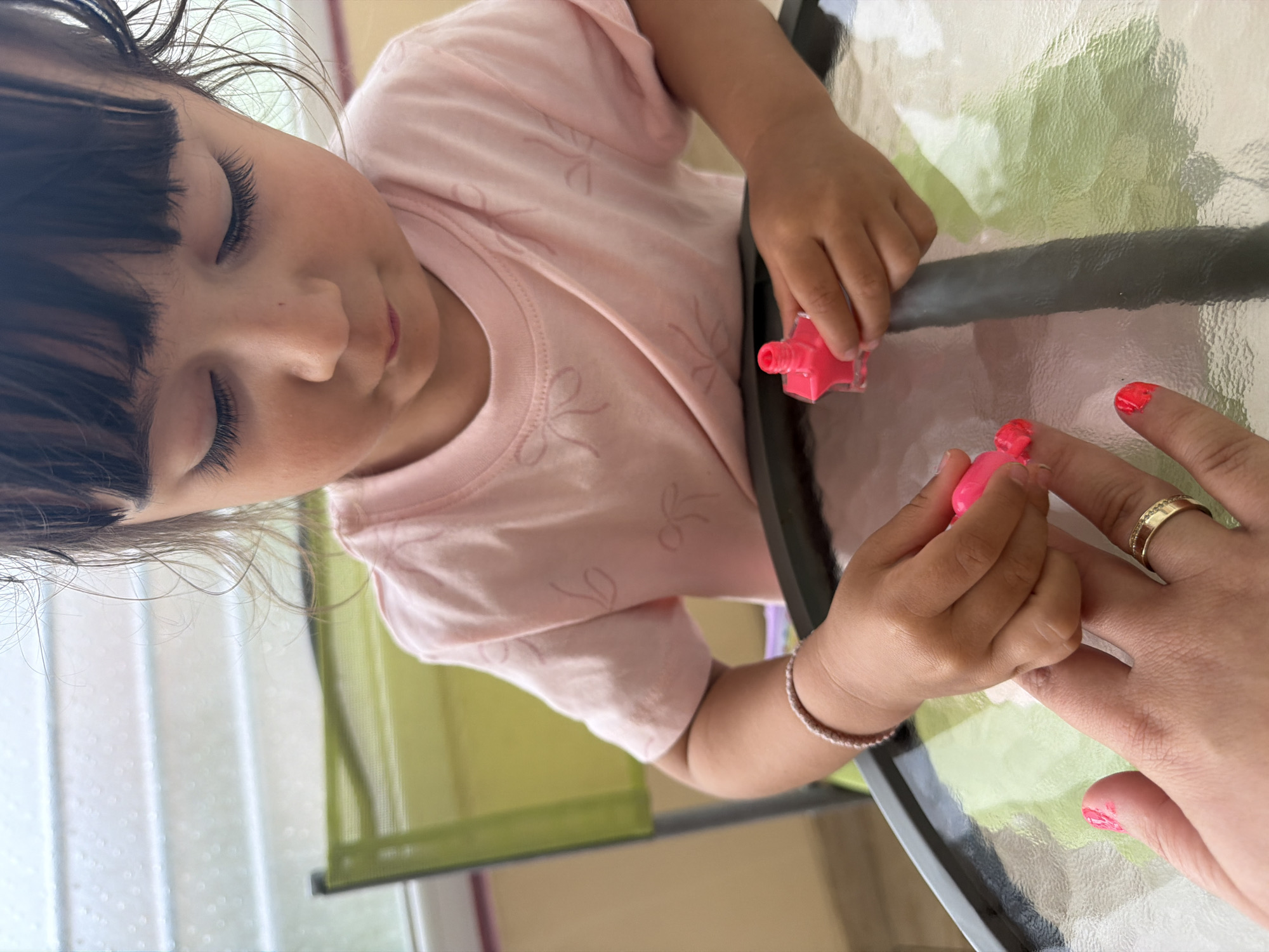 A young child, likely a girl, wearing a light pink t-shirt with subtle bow patterns, is engrossed in painting an adult's fingernails. The image is taken from a slightly overhead perspective, with the child positioned in the upper left, her face partially visible, showing closed eyes and dark, fringed bangs. Her expression suggests focus and concentration on the task.
The child holds a small, star-shaped bottle of bright pink nail polish in her right hand, while her left hand is poised over the adult's hand. One of the adult's fingers already has a vibrant red or pink polish applied, and there are smudges of the same color on another finger, indicating the ongoing activity. The adult wears a gold-toned ring with a textured or multi-band design on their ring finger. The child also has a light-colored beaded bracelet on her right wrist.
They are seated at a glass-top table that reflects parts of the surroundings, giving the surface a mottled green and white appearance. The background is softly blurred, showing hints of light-colored curtains or blinds and possibly outdoor greenery, suggesting an indoor or covered patio setting during daylight hours. The overall scene depicts a tender, playful moment between a child and an adult, possibly in Burgas, Bulgaria. No visible text is present in the image.