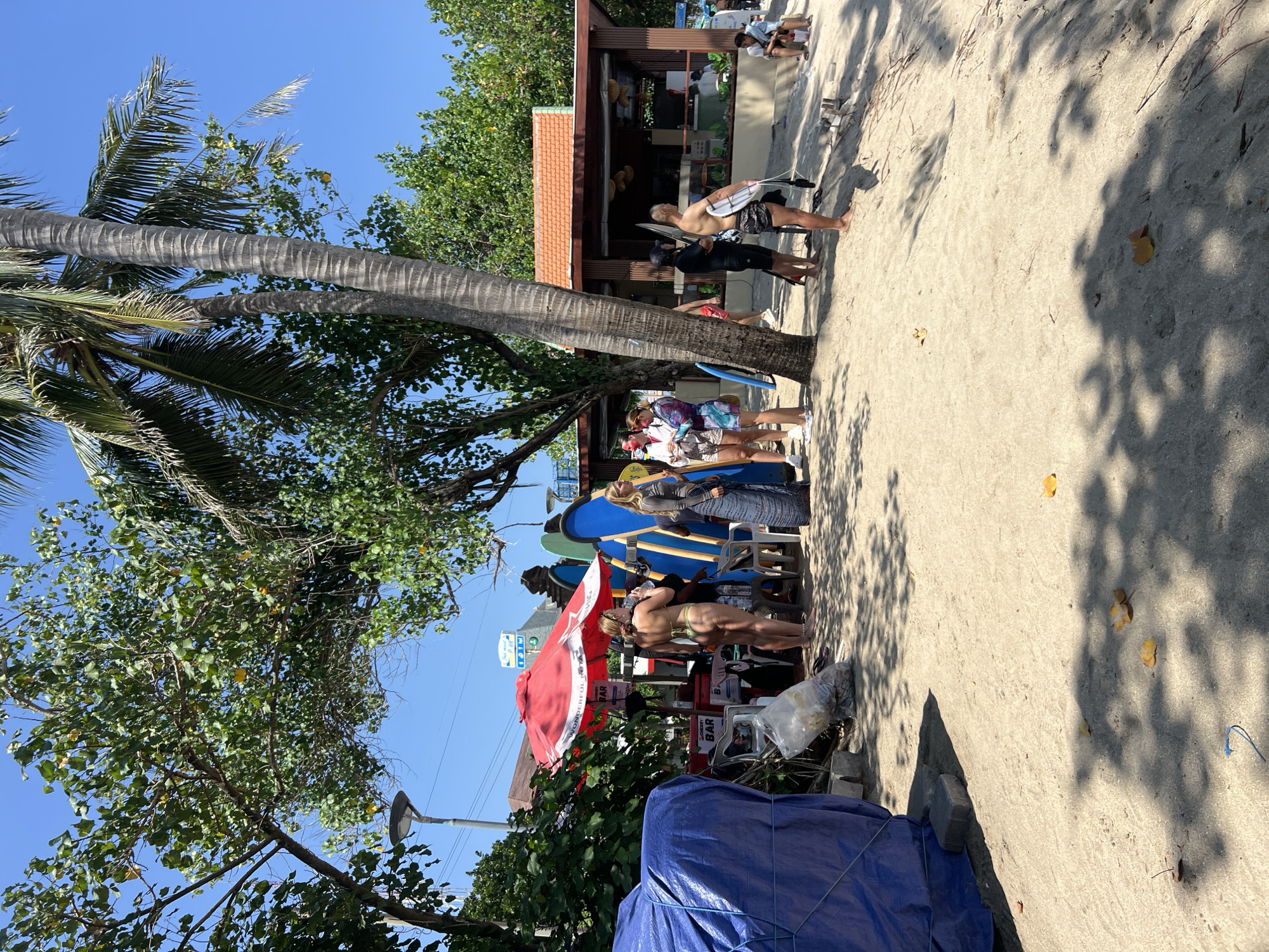 The image depicts a lively beach scene in Kuta, Indonesia, with a focus on a surf rental and bar area. In the foreground, the sandy beach is partially shaded by palm trees, with a few fallen leaves scattered across the sand. To the left, a large blue tarp covers an unknown object, with some discarded plastic bags and debris nearby.
The central area of the image features a row of surfboards, mostly blue, leaning against a structure that appears to be a bar. A bright red umbrella, labeled "WONDERFUL" and "BAR," shades the bar area. Several people are gathered around this area. A woman in a bikini stands near the bar, while others, including individuals in swimwear and casual clothing, are conversing or looking towards the ocean. Some individuals are holding surfboards.
Further to the right, a small building with a terracotta-tiled roof serves as a more established bar or shop. More people are visible in and around this building, some sitting and others standing. The background shows lush green trees and a clear blue sky. The overall impression is one of a popular and active beach destination.