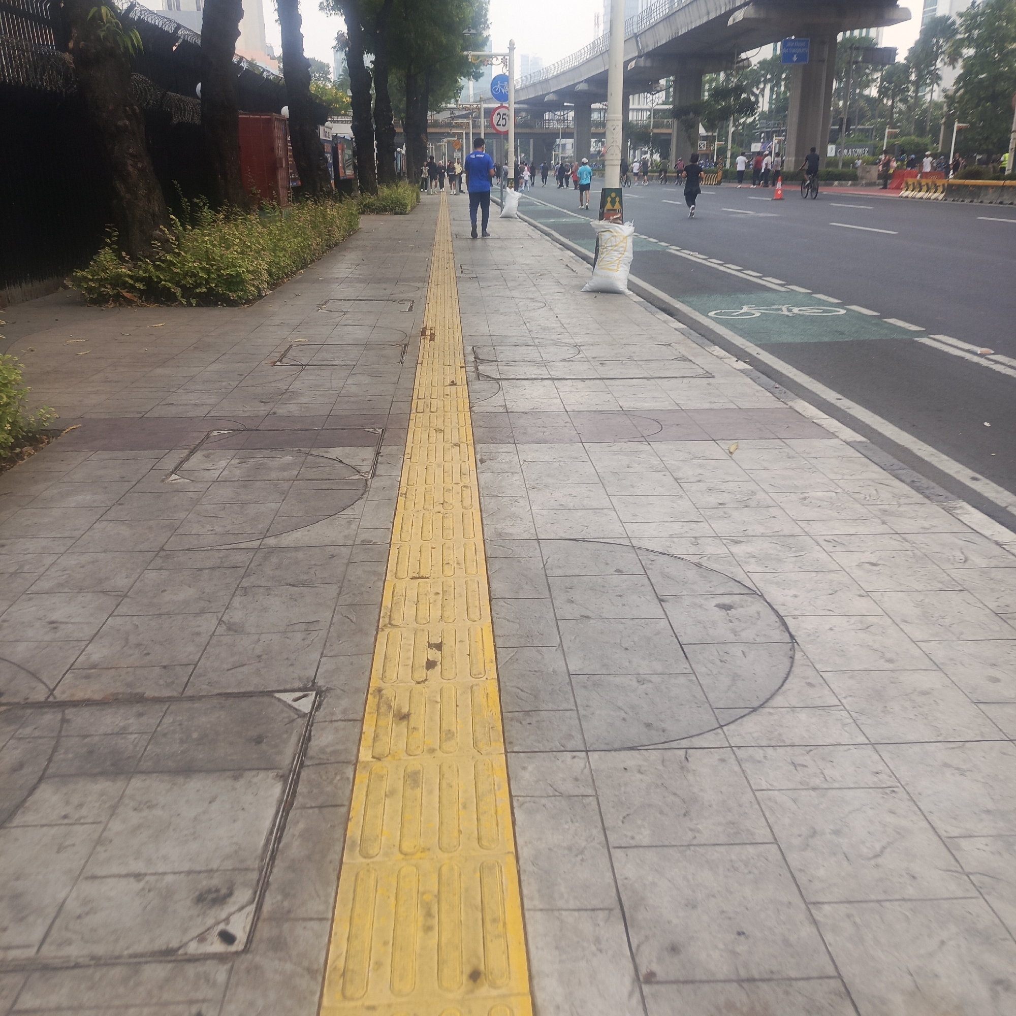 A busy street scene in South Jakarta, Indonesia, captures a pedestrian walkway alongside a busy road. The walkway is paved with gray tiles and features a bright yellow tactile paving strip, indicating it is a path for the visually impaired. Several people are walking on the sidewalk, some in athletic attire. The road itself has clear lane markings and a designated bicycle lane marked with green paint and a bicycle symbol. In the background, elevated infrastructure, possibly a highway or a mass transit line, looms over the scene. Trees line the edge of the sidewalk, providing some shade. The sky is overcast, suggesting a cloudy day. Several large white sacks, possibly containing sand or other materials, are placed along the edge of the walkway. There are also traffic signs visible, including one indicating a speed limit of 25. The overall impression is of an active urban environment with a focus on pedestrian and cyclist safety.