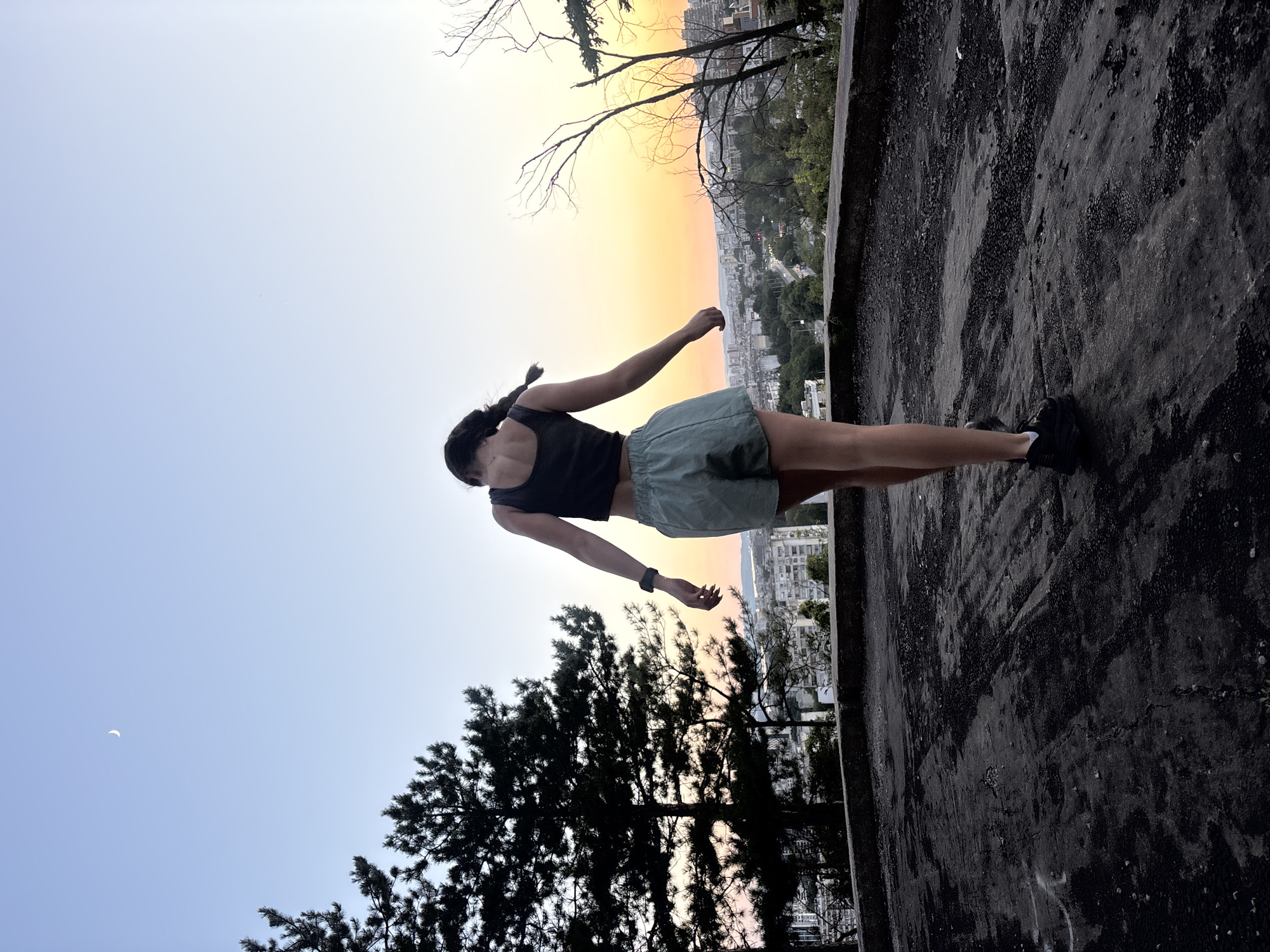 A young woman with dark hair braided down her back stands on a dark, textured rooftop, facing away from the camera and looking out over a city at sunset. She is wearing a dark sleeveless top and light green shorts. Her arms are slightly outstretched, and her head is tilted down. The sky is a gradient of pale blue to orange and yellow. To the left, a crescent moon is visible. Pine trees are silhouetted against the sky on the left side of the image, and bare branches of another tree are visible on the right. In the distance, the city of Varna, Bulgaria, is spread out with buildings and a highway visible. The rooftop appears to be concrete or asphalt, with some debris and markings. The overall scene suggests a moment of contemplation or observation at dusk.