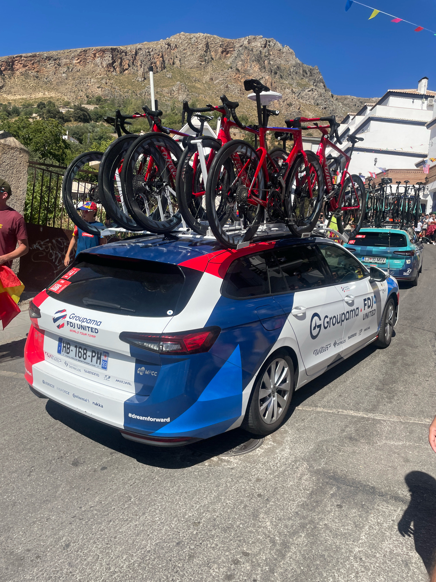 This is a medium-shot, eye-level, outdoor photograph taken on a sunny day in the city of Güéjar Sierra, Spain. The focus is on the rear of a white, blue, and red branded car, possibly a support vehicle for a cycling team, with multiple bicycles mounted on its roof rack. The car has "Groupama FDJ UNITED WORLD TOUR TEAM" branding on its rear door, and a license plate that reads "HB-168-PM 93". The car is parked on a paved street.
In the background, there are buildings with white facades and tiled roofs, typical of Andalusian architecture, cascading down a hillside. A rocky, barren mountain looms behind the buildings. The sky is clear and blue, with a few triangular flags strung across the top right of the frame, suggesting a local event or festival.
To the left of the car, a person wearing a maroon t-shirt and a dark baseball cap is partially visible. Another person, a young boy wearing a colorful cap, stands behind the car. Several other people are visible in the background, near a turquoise car parked behind the main subject. The scene appears to be part of a cycling race or event, given the presence of multiple bicycles and team branding.