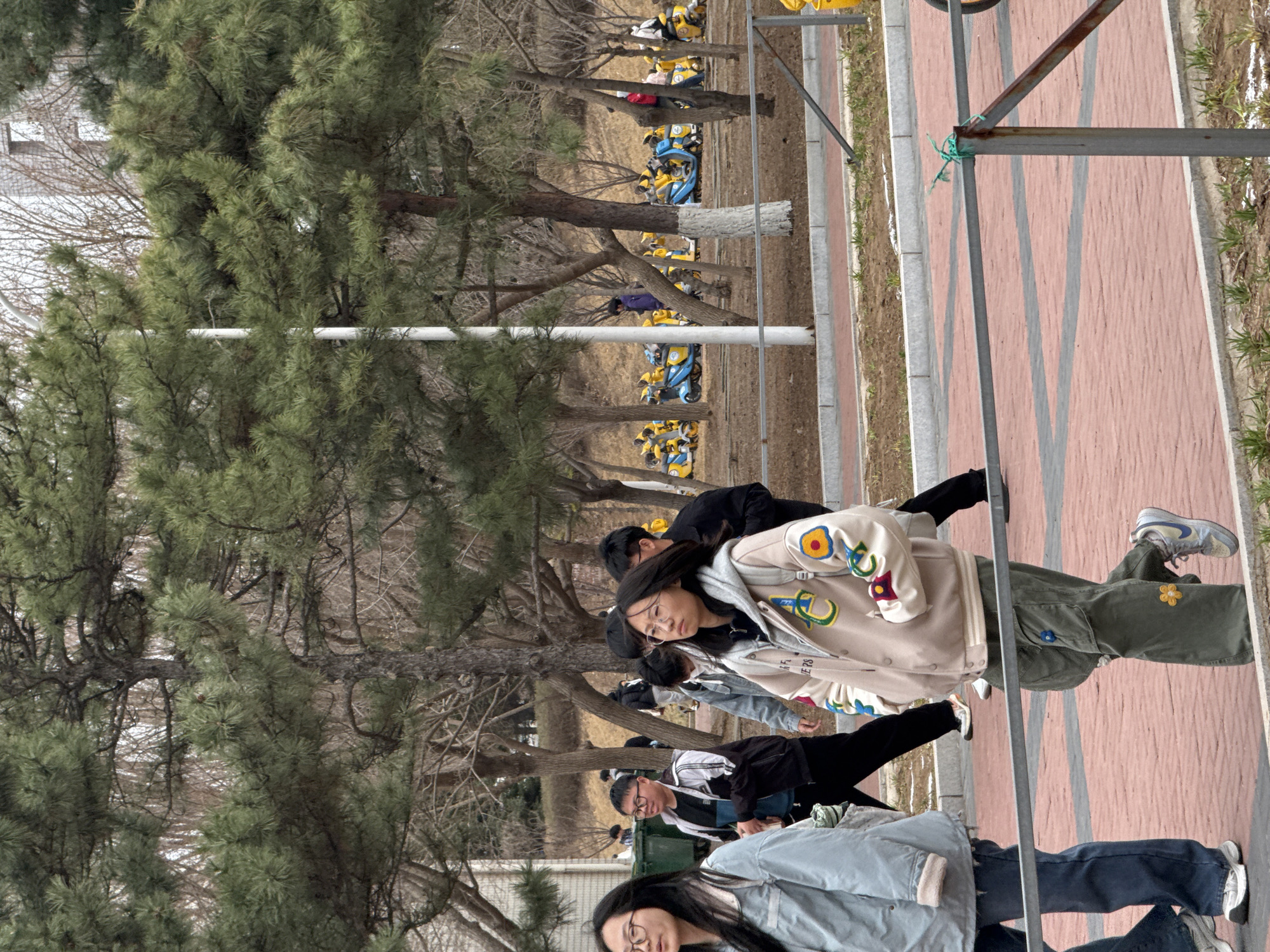 A group of people are walking along a brick path in Zhongtun, China. The path is lined with trees and a metal fence. In the background, there are rows of parked scooters. One person in the group is wearing a beige jacket with colorful patches and green pants with a yellow flower patch. Another person is wearing a blue jacket and jeans. The group appears to be walking towards the camera.