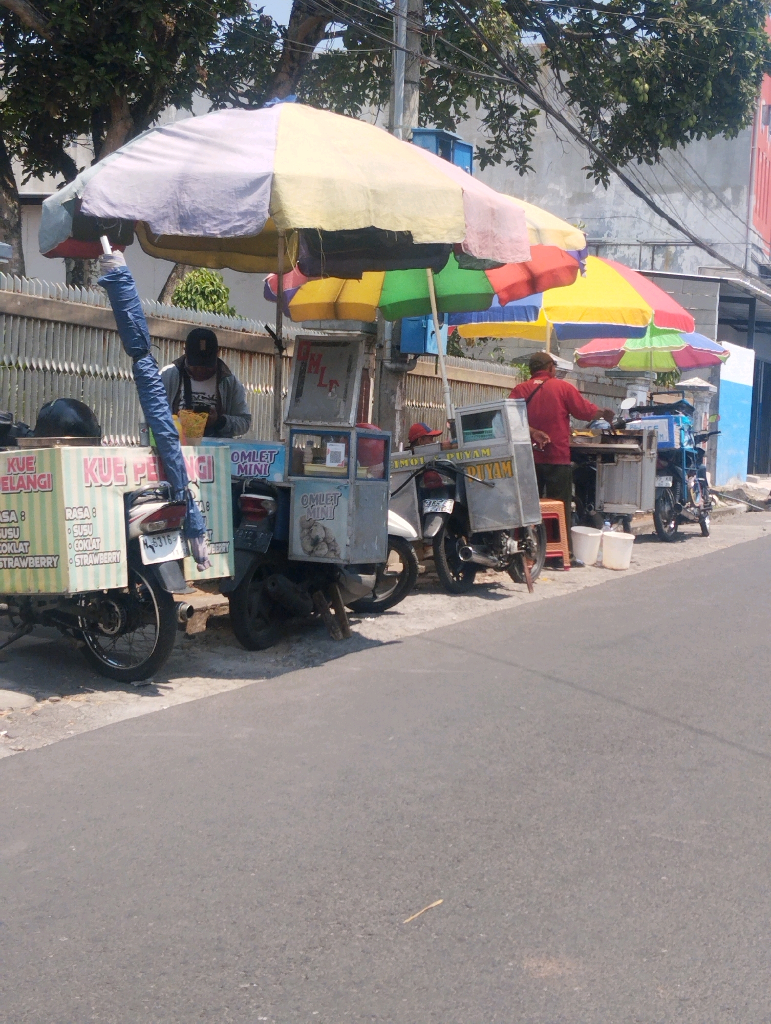 In Malang, Indonesia, on a sunny day, street food vendors are operating their businesses by the roadside. The scene features several food carts, each attached to a motorcycle. The carts are covered with colorful umbrellas, predominantly yellow, pink, and green.
One cart, labeled "KUE PELANGI," displays a variety of offerings, with text listing flavors like "RASA 8," "SUSU," "COKLAT," and "STRAWBERRY." A person wearing a dark cap and jacket is seen behind this cart, possibly attending to customers or preparing food.
Another cart, labeled "OMLET MINI," is also visible, with similar packaging and a similar setup. Further down the road, another vendor in a red shirt is actively cooking at their cart, with two white buckets placed nearby. The carts are parked on the side of a paved road, with a low metal fence behind them, and trees with green foliage providing shade. The overall environment suggests a typical urban street scene in Indonesia, with bustling street food commerce.
