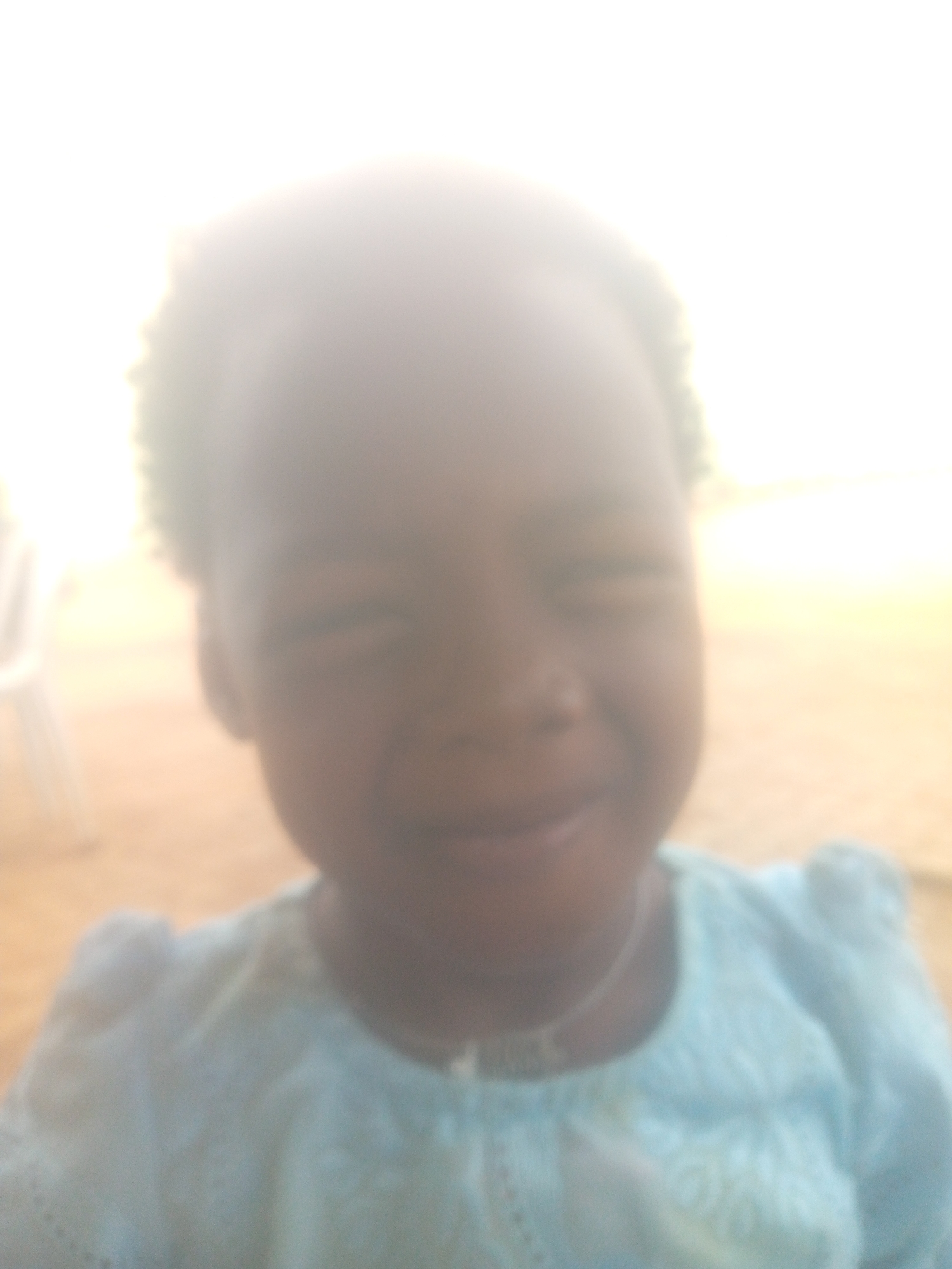 A severely overexposed and blurry close-up portrait shows a young child with dark skin and short, curly hair. The child is positioned centrally, looking towards the camera with eyes squinted, possibly from bright light or a shy smile, and a subtle grin. They are wearing a light blue top or dress, which appears to have a textured or patterned surface, possibly embroidered or knitted. A thin necklace with a small, indistinct pendant is visible around their neck.
The background is almost entirely washed out by extreme brightness, making specific features indiscernible. Hints of a light-colored object, possibly a chair, can be vaguely seen on the left side, and a light brown surface, likely the ground or floor, is present in the lower background. The overwhelming brightness suggests the photograph was taken during daylight, possibly outdoors or in a brightly lit indoor environment. Due to the significant overexposure and lack of focus, fine details of the child's features, clothing, and the surrounding environment are obscured. No legible text is visible in the image.