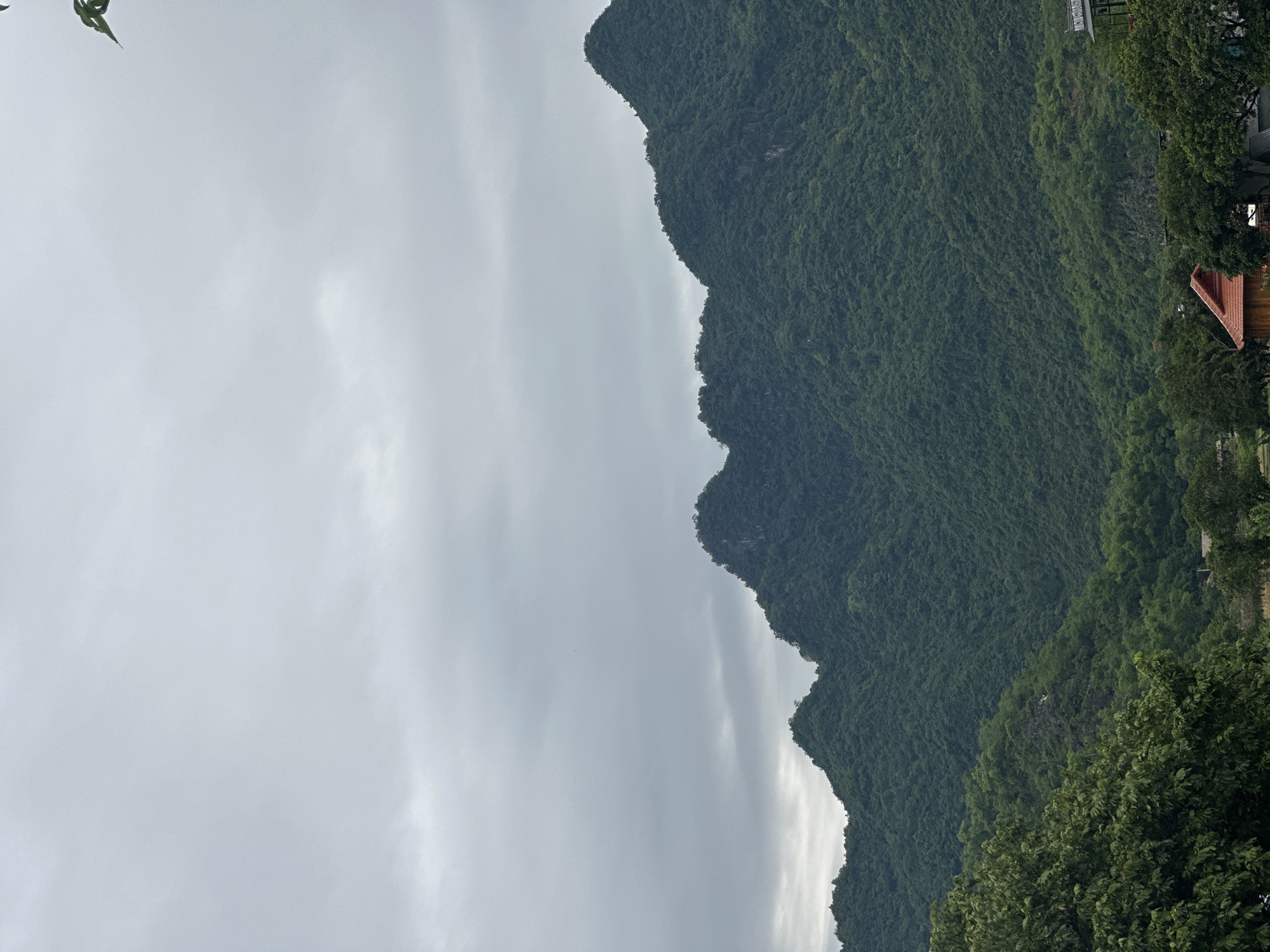 The image depicts a lush, green mountain range under a cloudy sky in Xóm Khán, Vietnam. The mountains are densely covered with vegetation, creating a textured, undulating surface. In the foreground, several trees with full canopies are visible, with their leaves appearing a deep green. Below the mountains and trees, a few buildings with red tile roofs and other structures are partially visible, suggesting a settlement or village. The overall atmosphere is serene and natural, with a hint of human habitation integrated into the landscape.