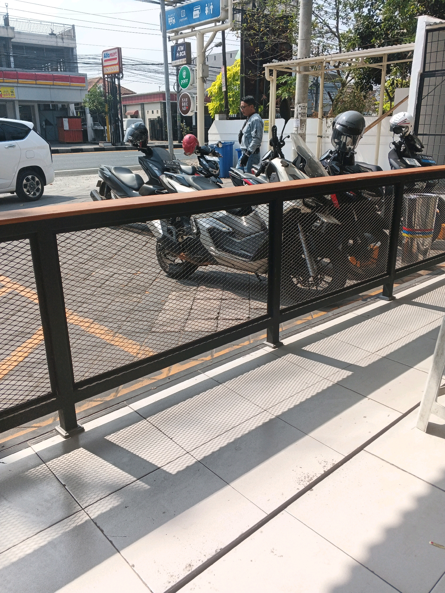 The image shows a street scene in Genuk Barat, Indonesia, with several motorcycles parked. In the foreground, a black metal fence with diamond-shaped mesh separates the viewer from the parked motorcycles. The motorcycles are parked on a paved surface with yellow markings. A white car is visible on the left side of the image, parked near a building that appears to be a convenience store, indicated by a red and white Alfamart sign. A man in a denim jacket and glasses is standing near the motorcycles, facing away from the camera. In the background, there are buildings, trees, and utility poles with wires. The lighting suggests it is daytime, with strong shadows cast on the ground.