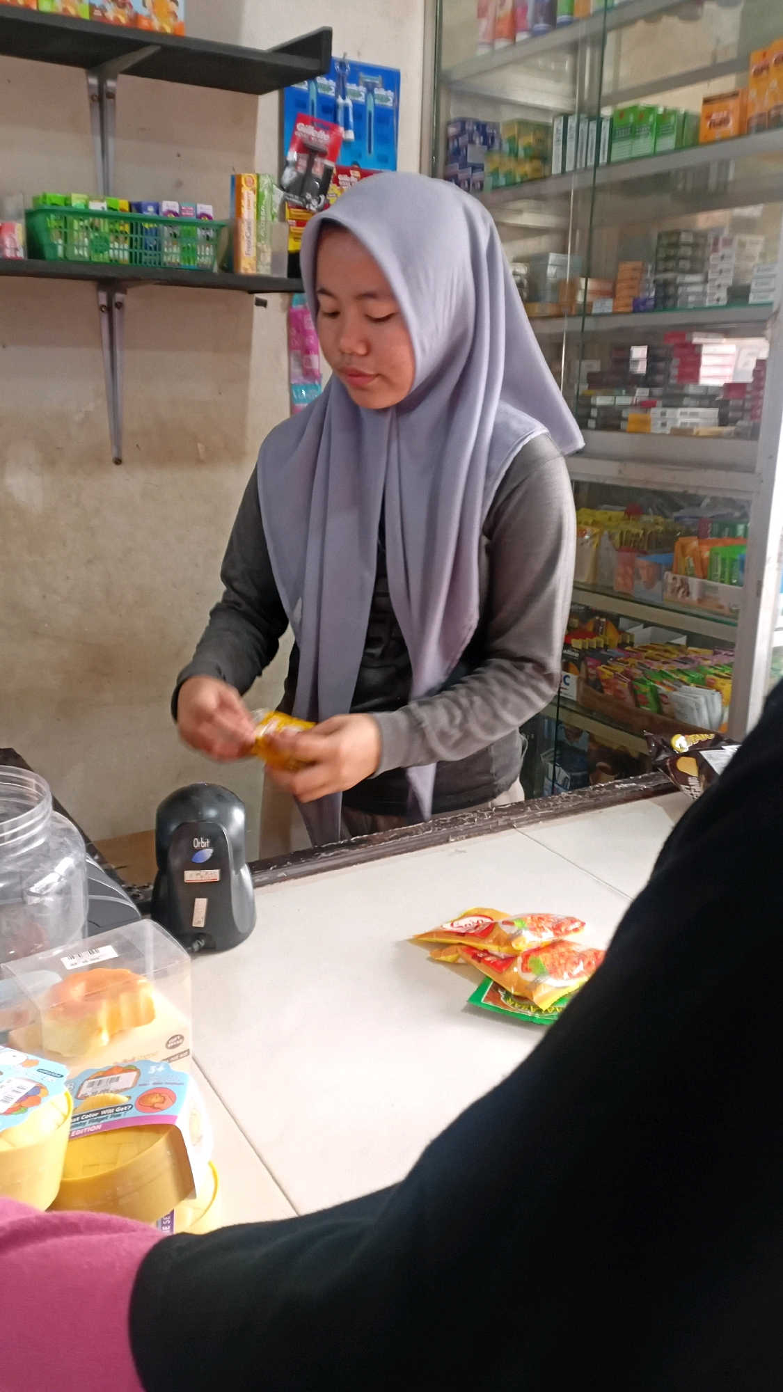 In Kasang Pudak, Indonesia, a young woman wearing a light purple hijab and a dark grey long-sleeved shirt is standing behind a white counter. She is holding a small yellow package in her hands, appearing to be opening it or examining its contents. In front of her, on the counter, is a black scanner device. To her left, a clear plastic jar is visible. Further to the right on the counter, several small, colorful packets are arranged, with one green packet underneath two yellow packets. Behind the woman, shelves filled with various packaged goods are organized within a glass display cabinet. The overall environment suggests a small retail store or convenience shop.