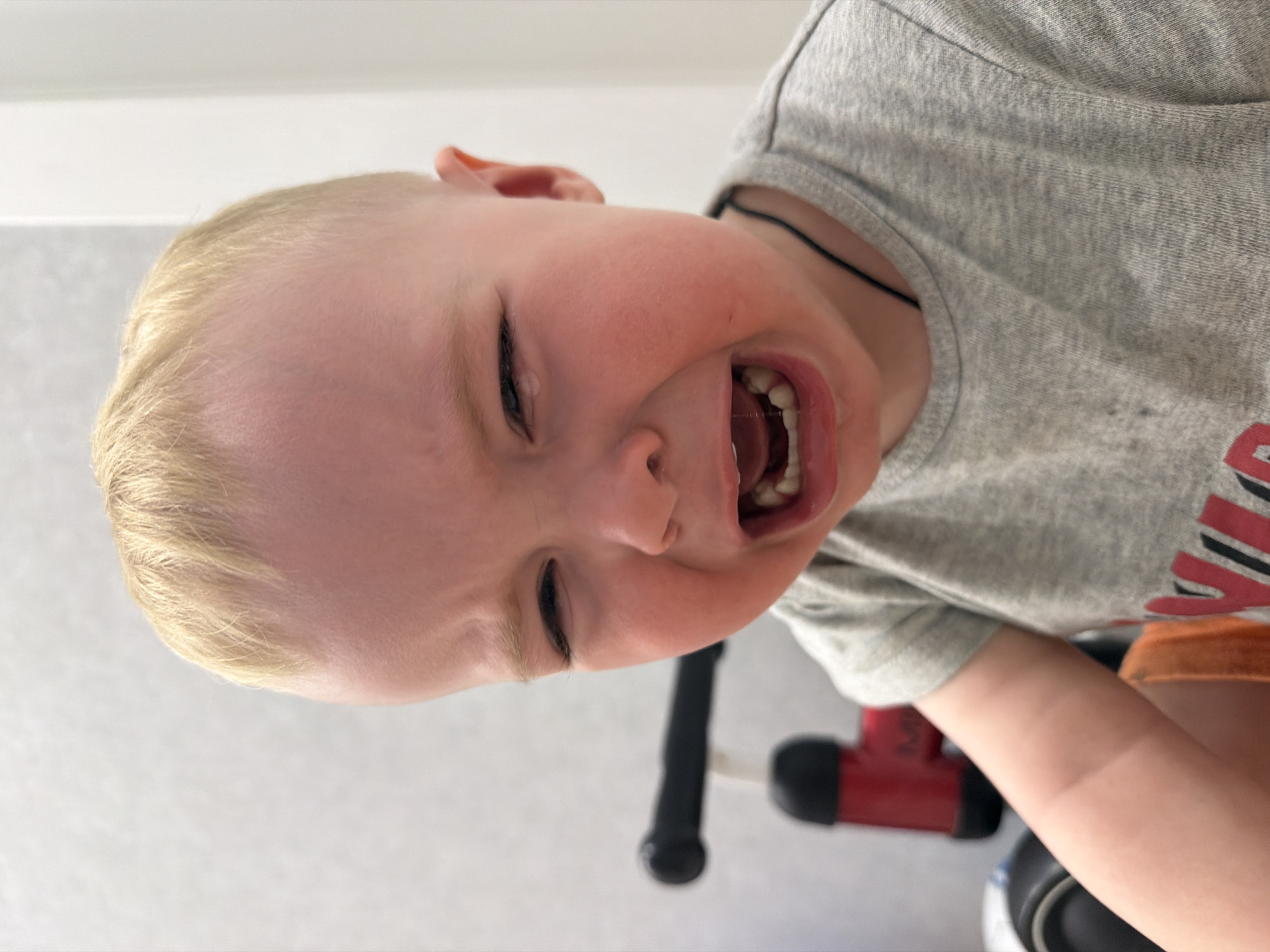 A blond-haired toddler is captured mid-cry, his face contorted with distress and his mouth open, revealing his teeth. He is wearing a gray T-shirt with a black neck cord. The image is taken from a low angle, showing part of a red and black toy vehicle in the background. The backdrop is a plain, light-colored wall. There is no evidence to confirm the image was taken in Votkinsk, Russia.