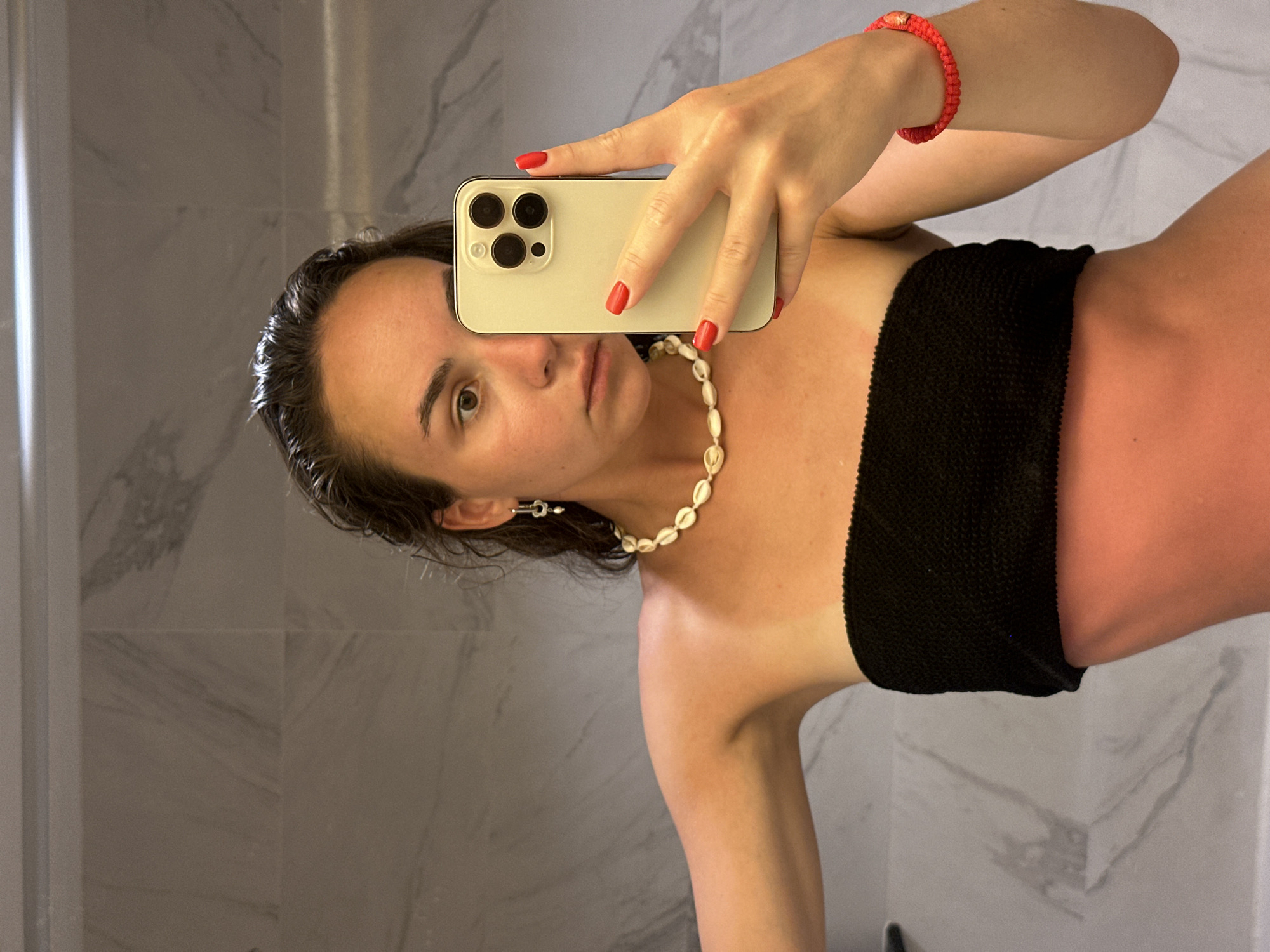 A young woman with wet dark hair and red nail polish is taking a selfie in a bathroom with light gray marble-like tiles. She is holding a white iPhone with three lenses, partially obscuring her face. She is wearing a black strapless top and a seashell necklace. A red beaded bracelet is on her right wrist. Her left ear has a small silver-colored earring. She is likely in Pattaya City, Thailand. Her skin on her right shoulder, chest, and upper abdomen is red, potentially indicating sunburn.