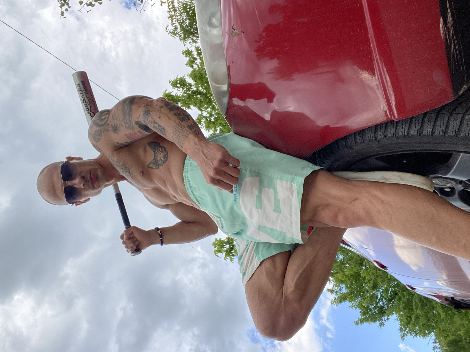 An adult male with a shaved head and sunglasses is positioned outdoors near a red vehicle. He is shirtless, wearing light-colored patterned shorts, and holding a baseball bat. Tattoos are visible on his torso and arms. The scene includes the vehicle's tire, wheel well, and body. The background shows trees and a partly cloudy sky. The location in the city of Pleven, Bulgaria, is not discernible from the image.