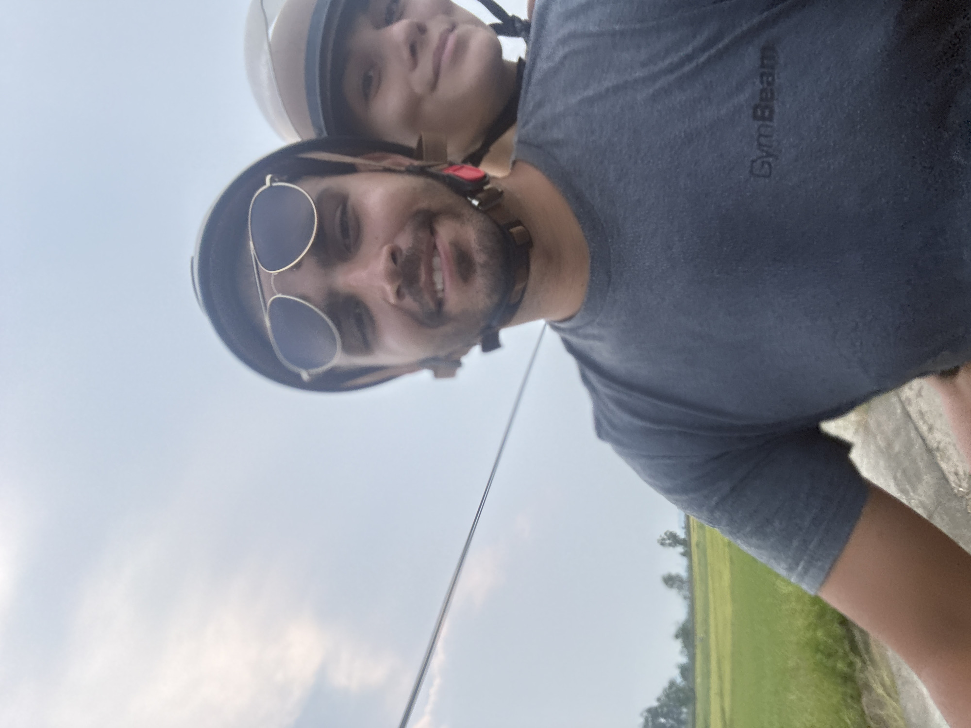 The image, rotated 90 degrees clockwise, captures a selfie of two individuals, presumably a couple, engaged in an outdoor activity in Đà Nẵng, Vietnam. The photo is taken from a low angle, with a bright, partially cloudy sky forming the dominant background.
In the foreground, a man with a beard, wearing dark aviator sunglasses and a dark brown helmet, smiles broadly at the camera, conveying happiness. He is dressed in a dark grey t-shirt with the brand name "GymBeam" visibly printed on the chest. Positioned behind him, a woman wearing a lighter beige helmet looks towards the camera with a neutral to slight smile. Both individuals wearing helmets strongly suggests they are riding a motorcycle or scooter.
The distant background reveals expansive green fields and some foliage, characteristic of rural or semi-rural landscapes often found outside urban centers in Vietnam. A thin, dark cable or wire extends horizontally across the middle of the sky. The scene indicates a daytime setting with clear weather, suggesting a pleasant journey or outing.