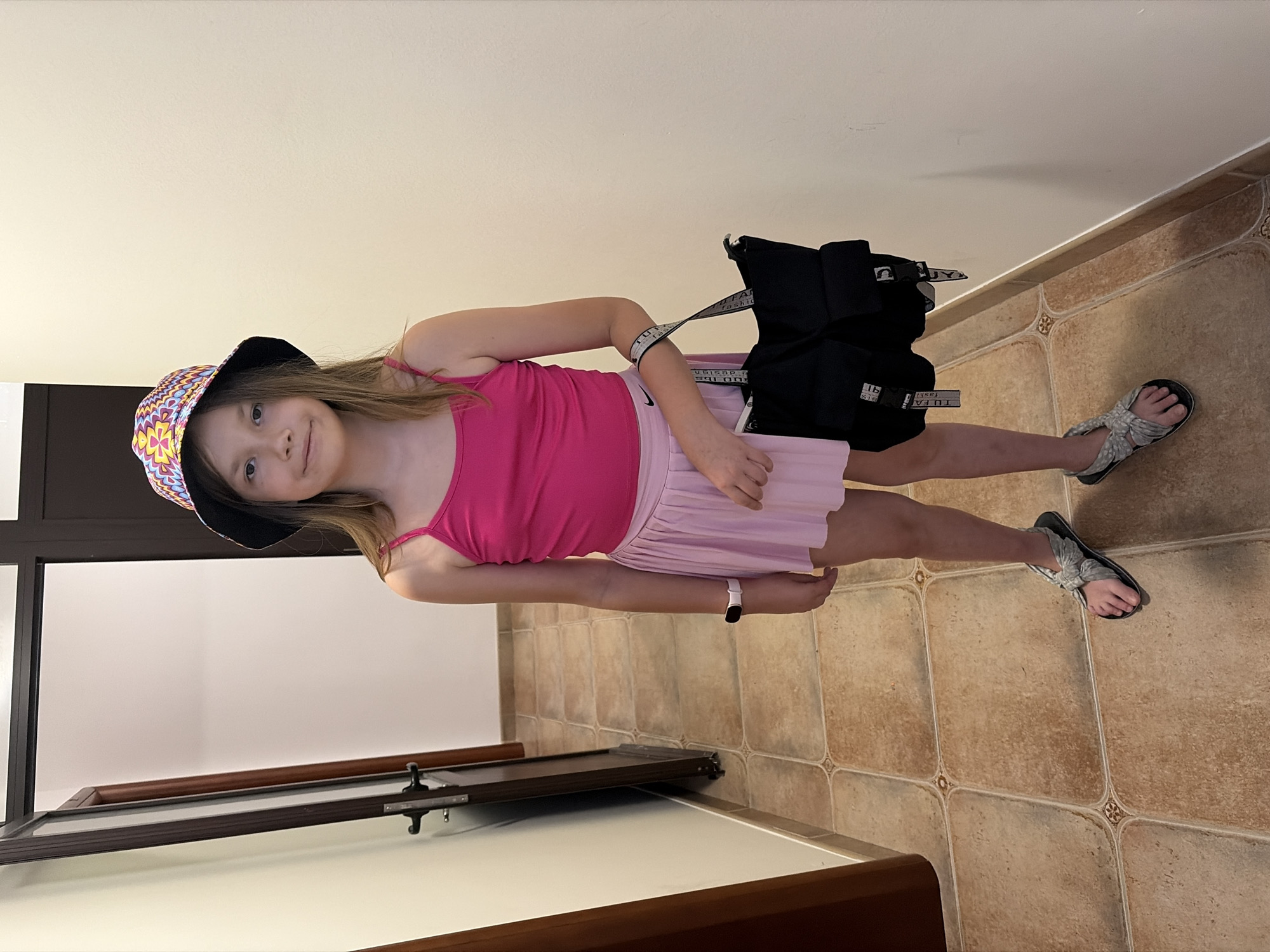 A young girl stands in a room. She wears a pink tank top, a light pink skirt, and patterned bucket hat. She has a black backpack slung over her shoulder. She wears sandals on her feet. The room has beige tiled flooring and a white ceiling. A dark-colored window frame is partially visible on the left.
There is no evidence in the image to determine its location in the city of Budva, Montenegro.