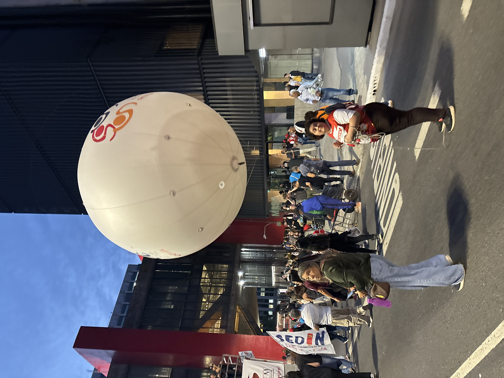 The image depicts a street scene in São Paulo, Brazil, with a large white balloon emblazoned with colorful logos hovering near a modern building with steel beams and glass windows. A crowd of people, mostly facing the same direction, are present. Some are seated, others standing. Several individuals wear masks. One person, closer to the camera, is walking along the street carrying a white balloon. A banner with the word "SEDIN" is visible. The sky above the building is a light blue dusk. A street with white markings is visible.