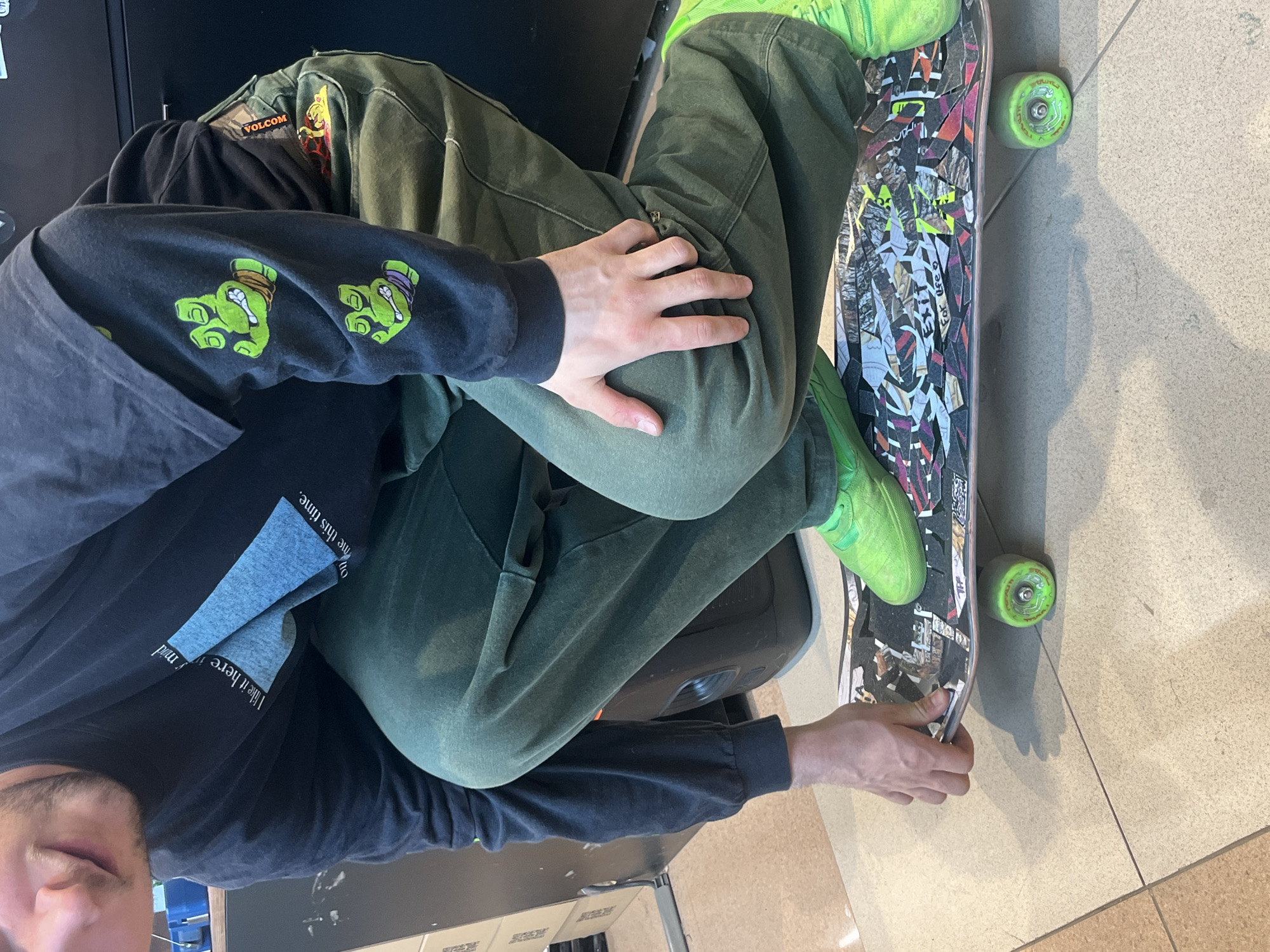 A man is sitting in a chair with a skateboard. He is wearing green shoes, green pants, a long-sleeved black shirt, and a black t-shirt. The skateboard has green wheels and is covered in stickers. The floor is tiled. The exact location in Krasnoyarsk, Russia, is not discernible from the image.