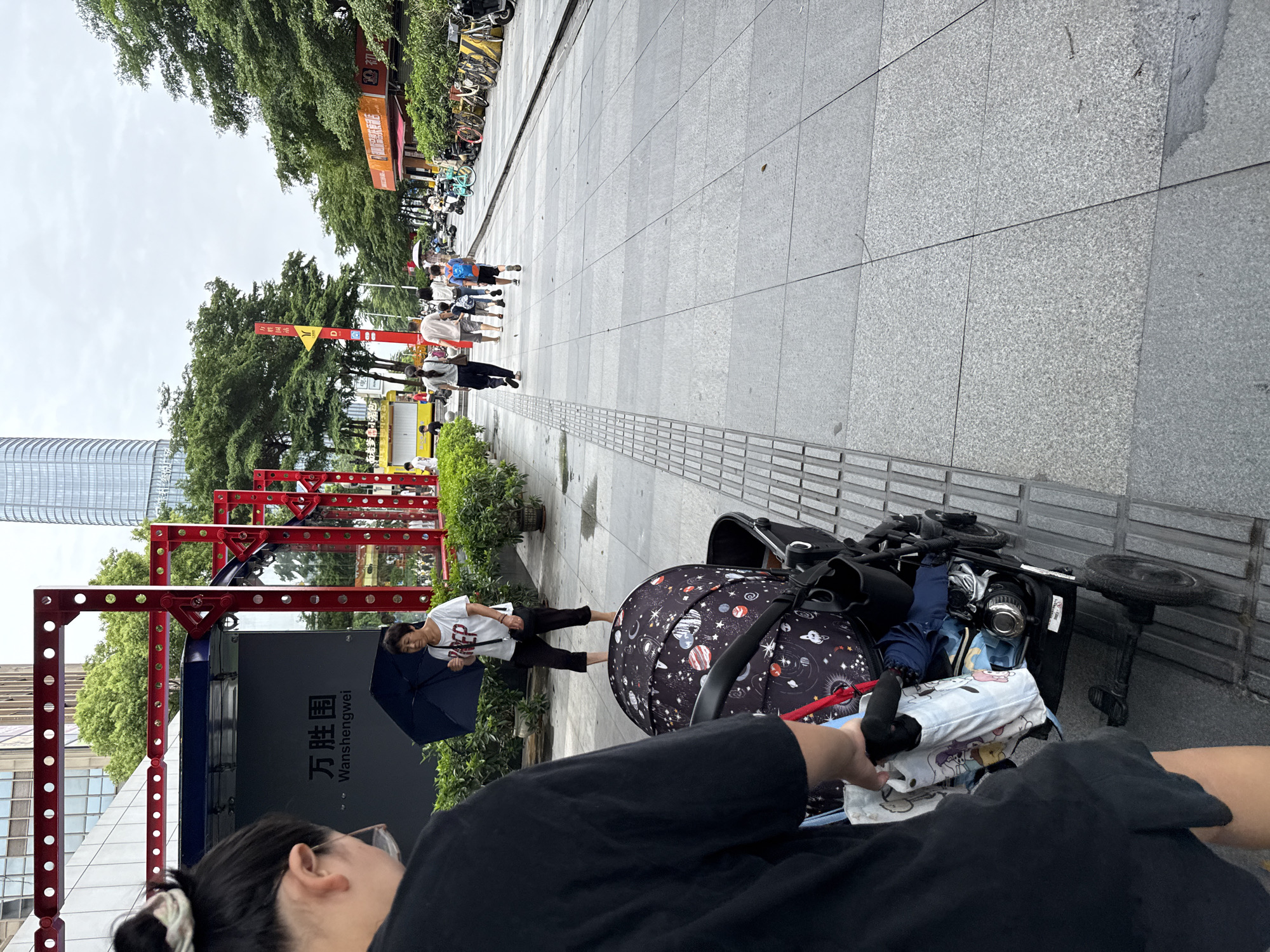 In Haizhu District, China, a person wearing a black t-shirt and shorts stands in the foreground, facing away from the camera and pushing a black stroller with a colorful galaxy-themed cover. The stroller is loaded with various items, including a white bag with cartoon characters. To the left, a woman in a white t-shirt and black cropped pants walks under a dark umbrella. Behind her, a red metal structure with circular cutouts is visible, alongside lush green bushes. Further down a paved walkway, a group of people, including children, are walking. The background features trees, buildings, and a bright yellow bus. The ground is a light gray tiled surface.