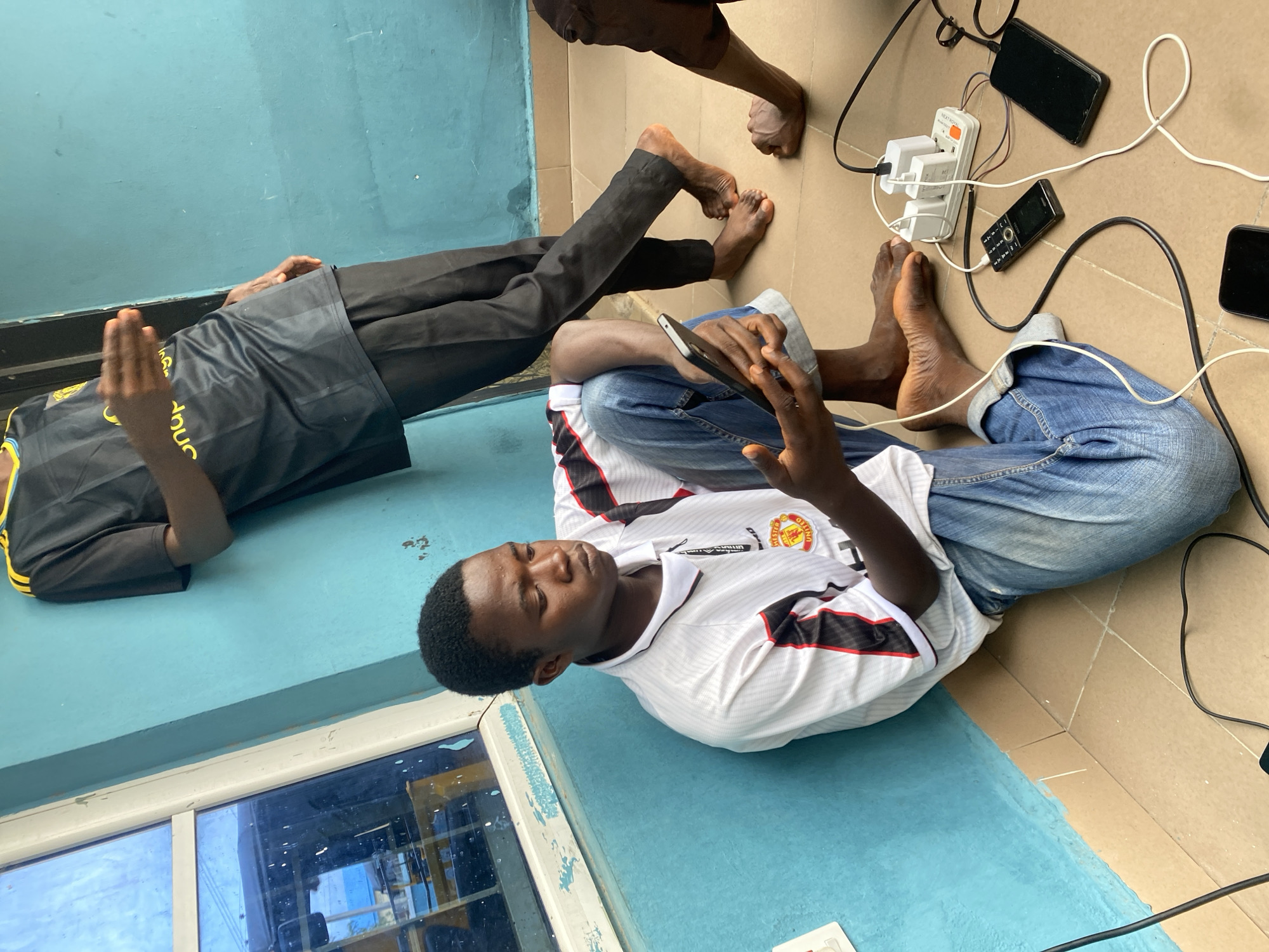 In Jalingo, Nigeria, three young men are gathered near a bright blue wall. One man is seated on the tiled floor, leaning against the wall, engrossed in his smartphone. He wears a white Manchester United jersey with red and black accents, and blue jeans. Another man stands behind him, also leaning against the wall, wearing a black and yellow striped athletic shirt and black pants, his hand gesturing outwards. A third person's legs are visible in the foreground, and part of their arm is seen on the right. Several electronic devices, including mobile phones and a power strip with multiple plugs, are scattered on the floor, connected by white and black charging cables, indicating they are charging. A window with blue-tinted glass is to the left of the frame. The scene appears to be an outdoor or semi-outdoor space, possibly a courtyard or balcony.